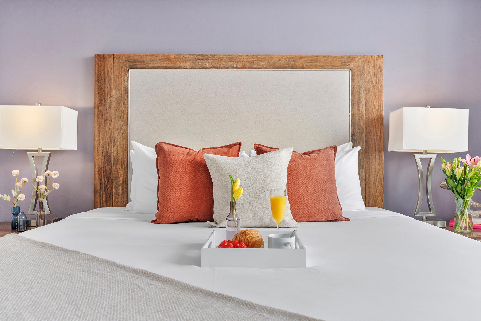 A head-on view of the king-sized bed with a large wooden and upholstered headboard, adorned with white and rust-colored pillows. Two nightstands with lamps flank the bed, and a breakfast tray is centered on the duvet.