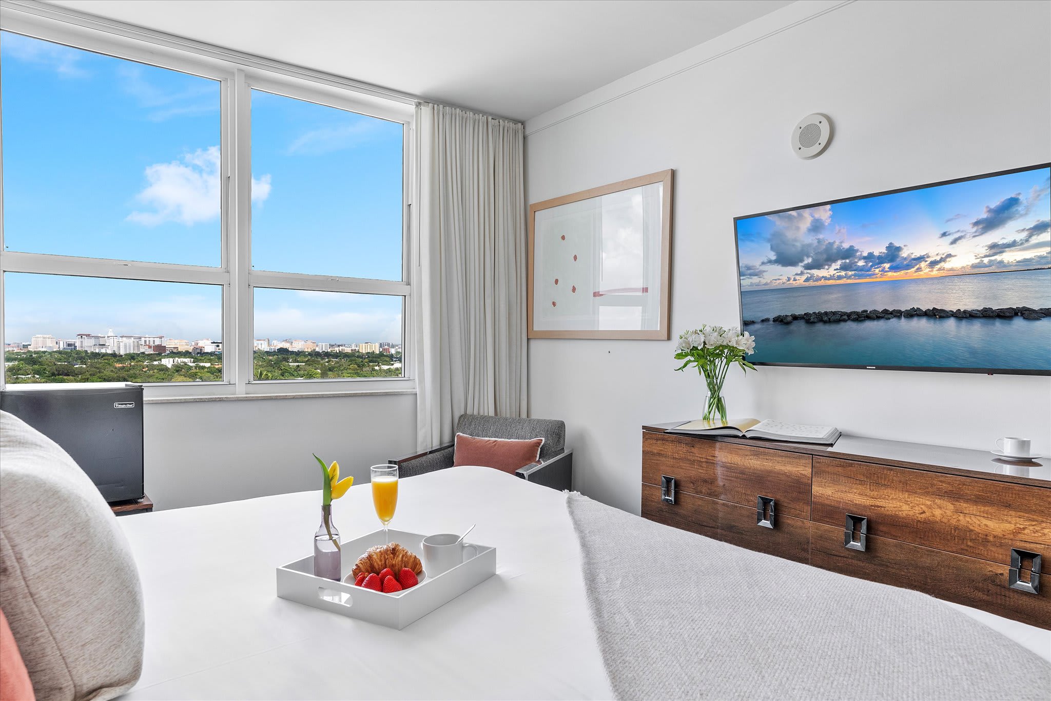 A bright bedroom featuring a white bed with a grey throw and a breakfast tray, positioned near a large window offering a city view. A flat-screen TV is mounted on the wall above a wooden dresser.