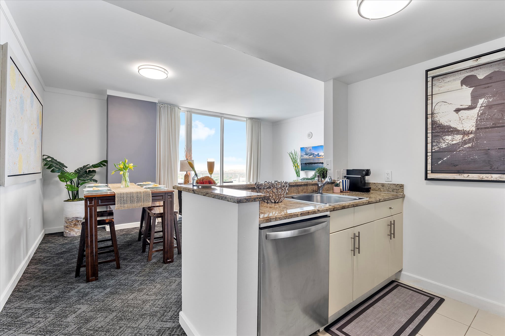 A view of the kitchen counter and dining area, showcasing granite countertops, a stainless steel dishwasher, and a breakfast bar. The wooden dining table and a glimpse of the living area and balcony view are visible in the background.
