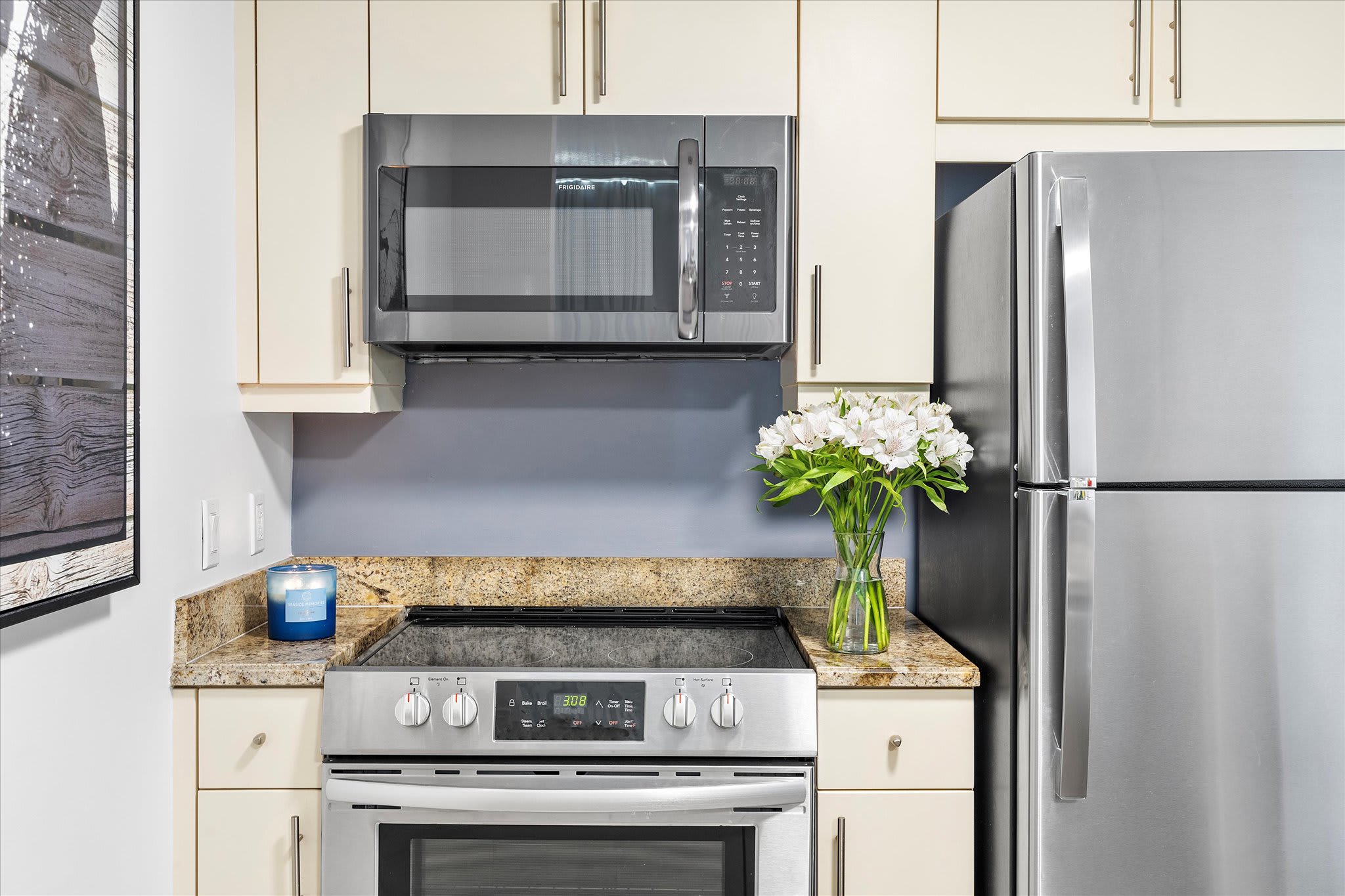 A close-up of the kitchen shows a stainless steel electric range with an overhead microwave, light-colored cabinets, and granite countertops. A vase with white flowers and a blue candle add decorative touches to the space.