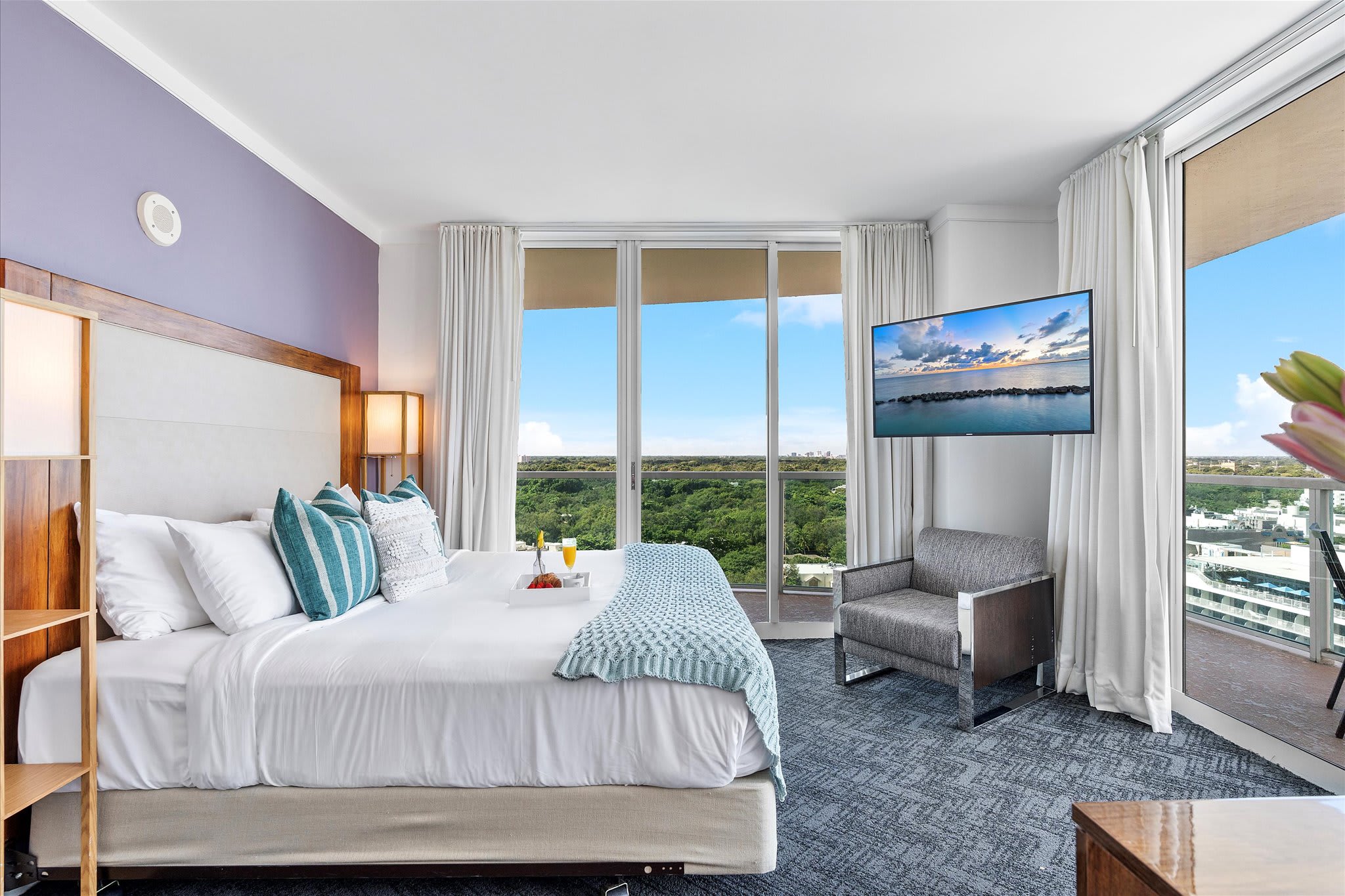 A bright bedroom with a large bed, light purple accent wall, and floor-to-ceiling glass doors leading to a balcony with a view of lush greenery and distant water. A wall-mounted TV and a grey armchair are visible.