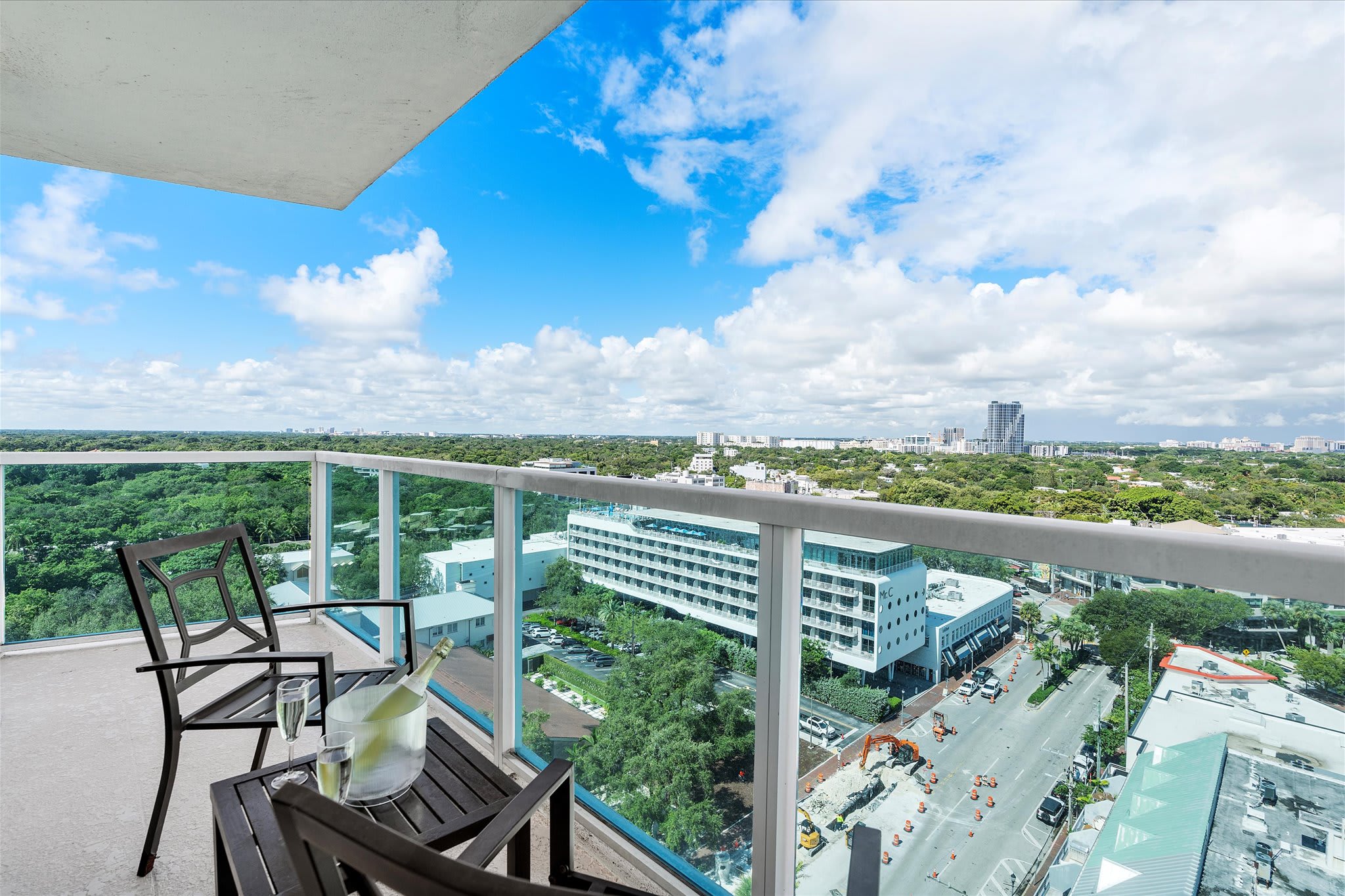 A high-rise balcony with glass railings, furnished with two black chairs and a small table holding a bottle of champagne and glasses. It offers an expansive view of the city, lush green areas, and distant water under a blue sky.