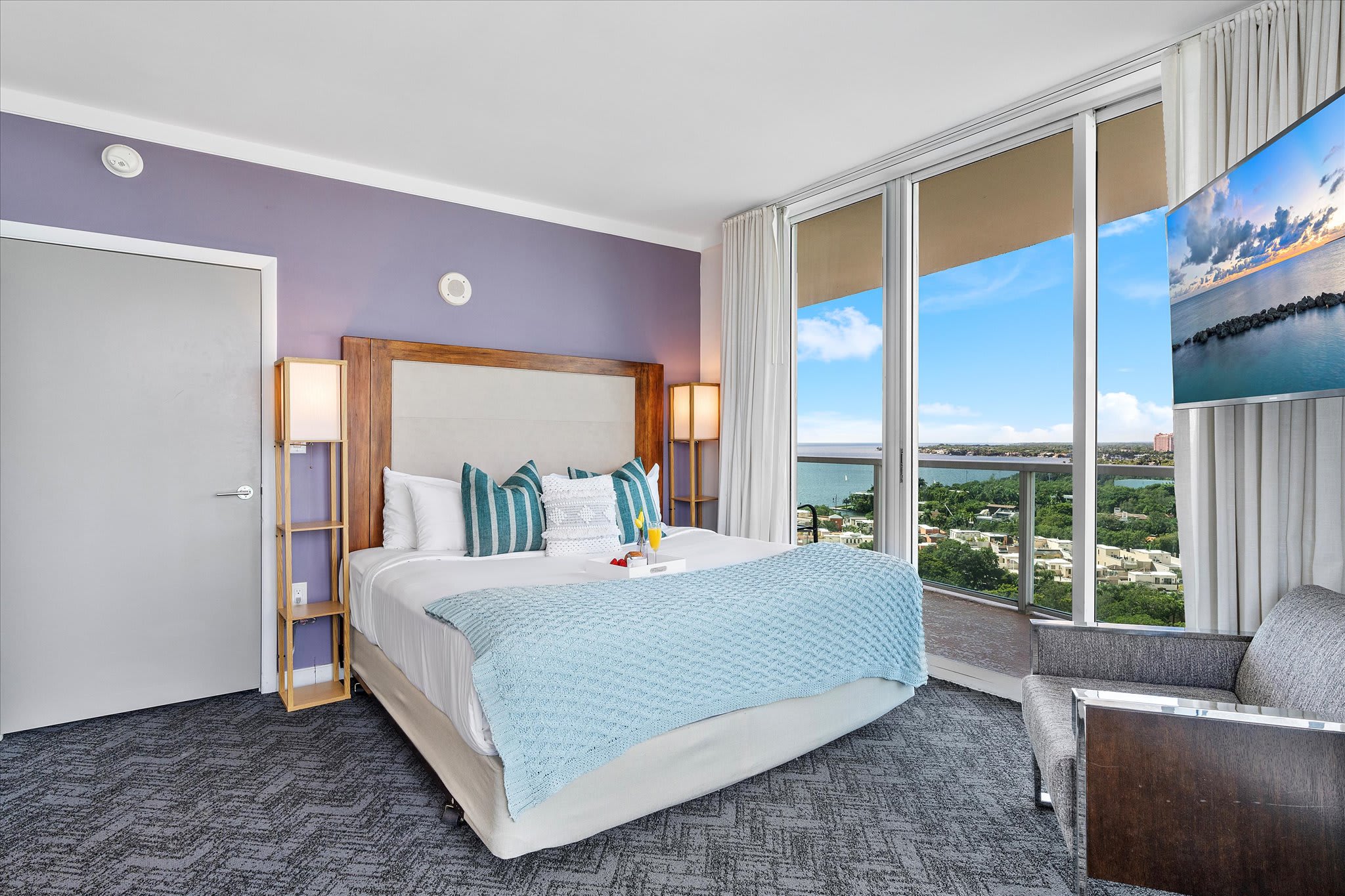 A spacious bedroom featuring a large bed with teal and white pillows, a light purple accent wall, and a full-length glass door opening to a balcony with a city and water view. A closed door to the left suggests an entrance or closet.
