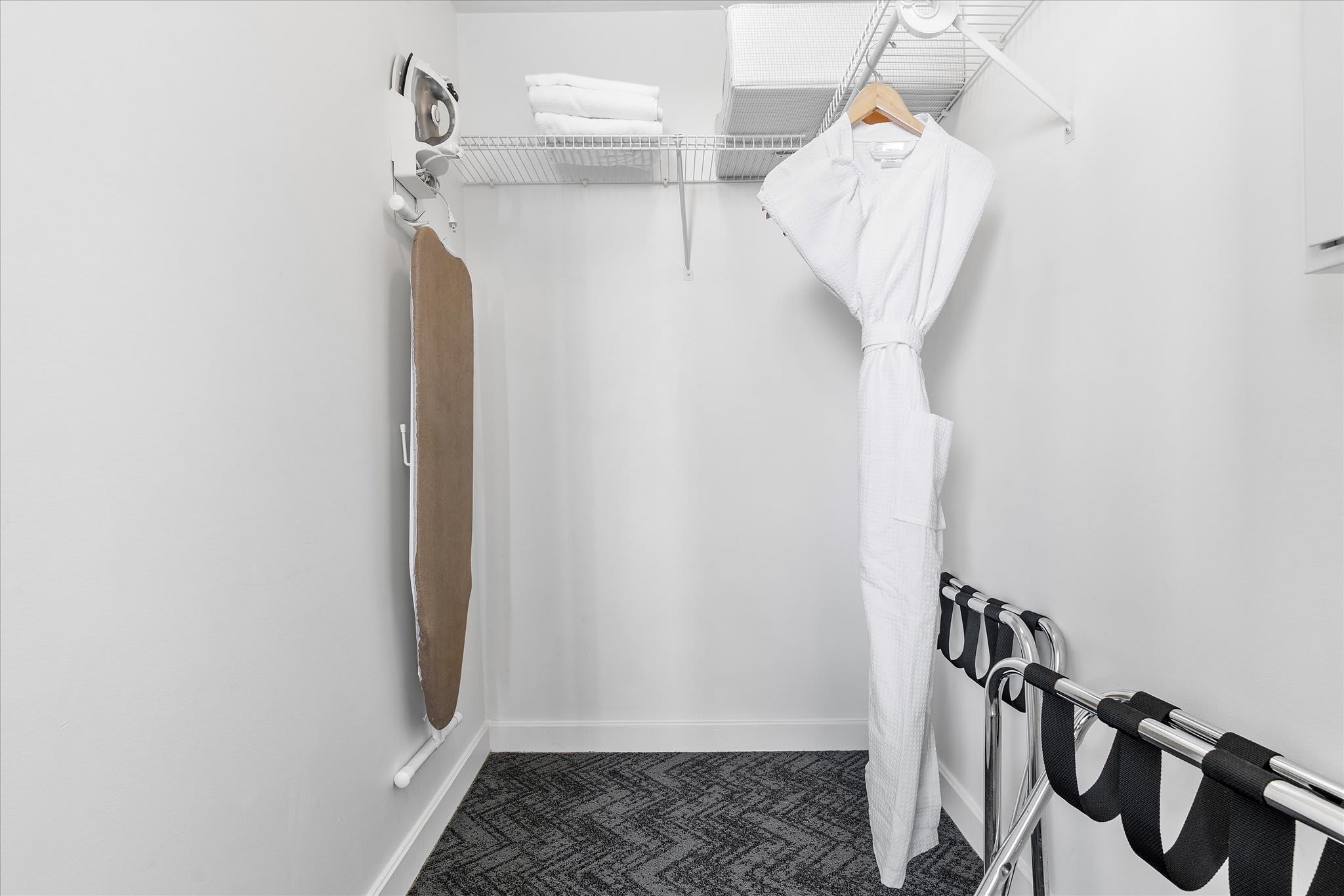 A walk-in closet with white wire shelving, an ironing board, and a white bathrobe hanging. The floor is covered with the same patterned grey carpet as the bedroom.