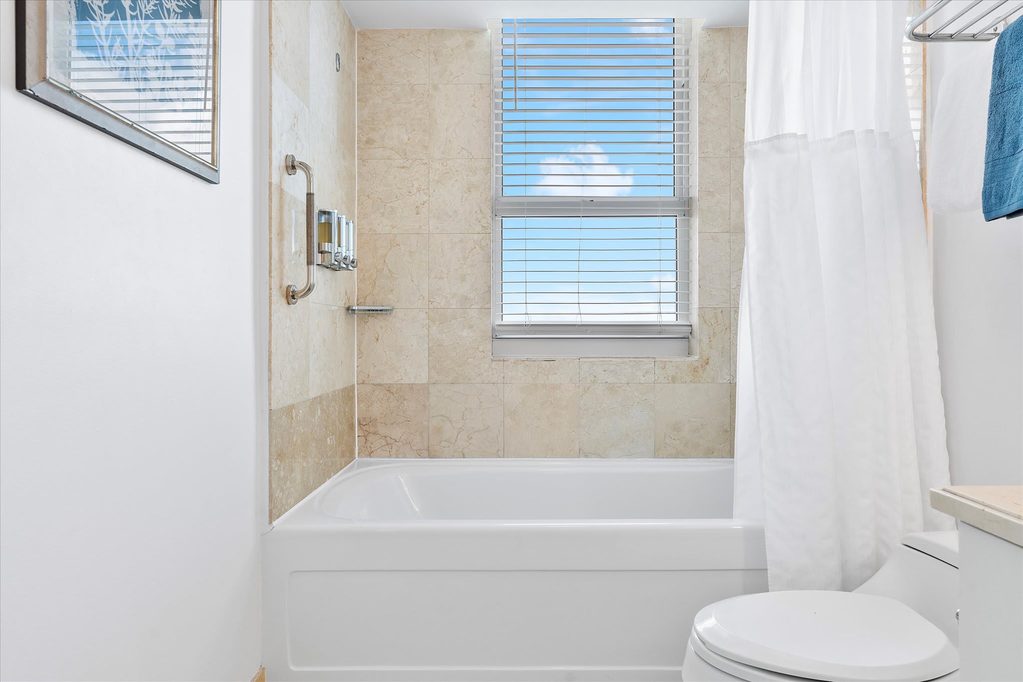 A close-up of the bathroom's white bathtub and beige tiled walls, featuring a window with white blinds. A shower curtain is pulled back, and wall-mounted dispensers are visible.
