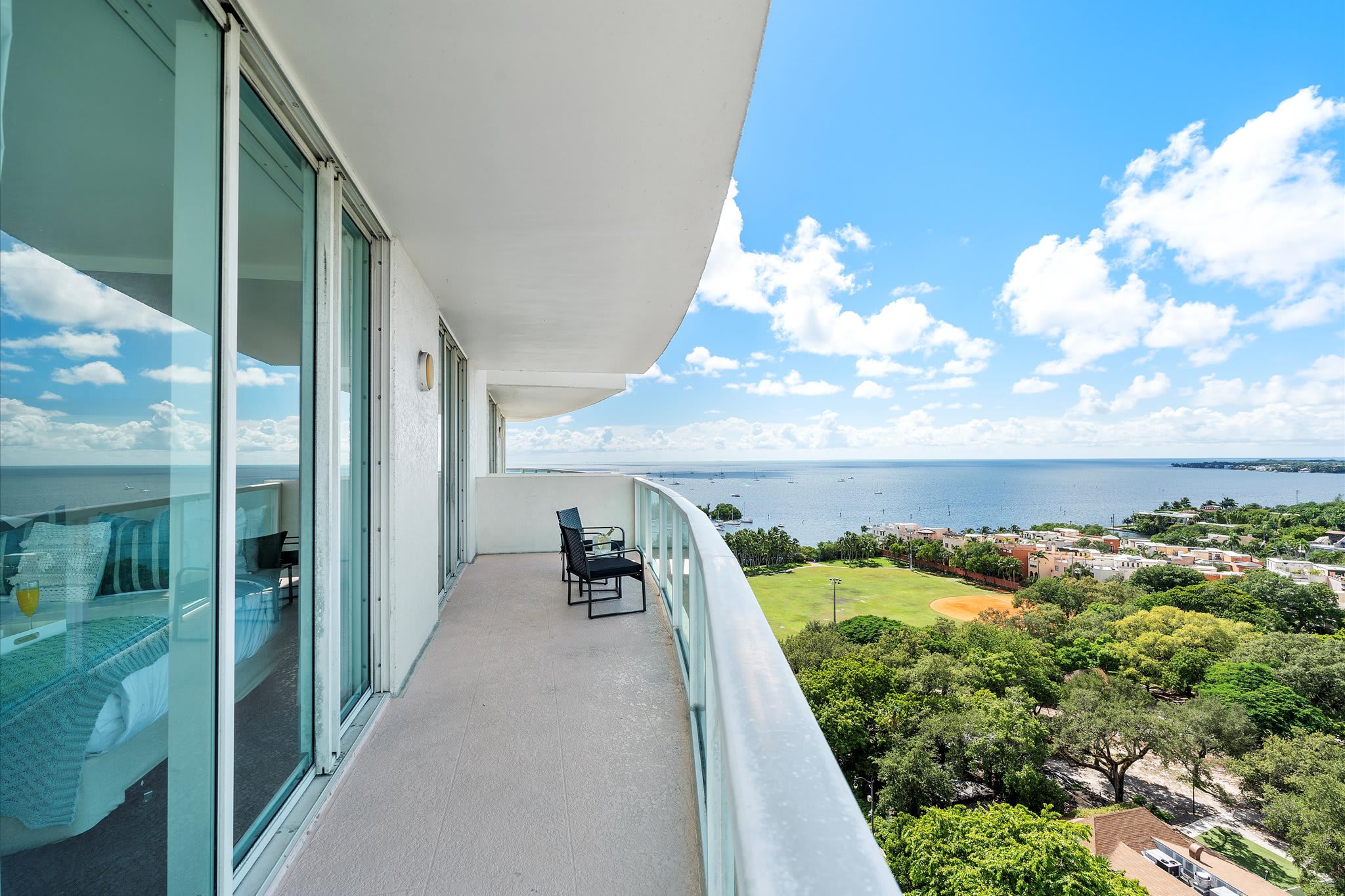 A spacious curved balcony with a glass railing offers panoramic views of the expansive bay, lush green trees, and a baseball field below. Two black outdoor chairs provide seating to enjoy the scenery.