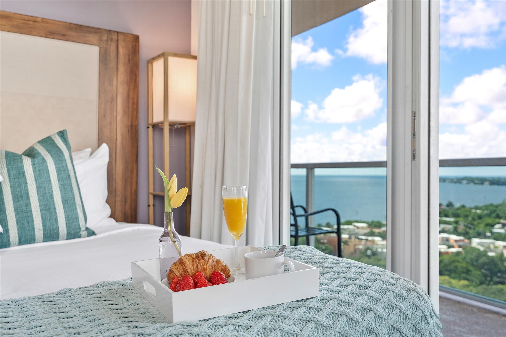 A close-up of a white bed with a light blue knitted throw, featuring a white breakfast tray with a croissant, strawberries, orange juice, and a coffee cup. A yellow tulip in a vase and a partial bay view through the balcony doors are visible.