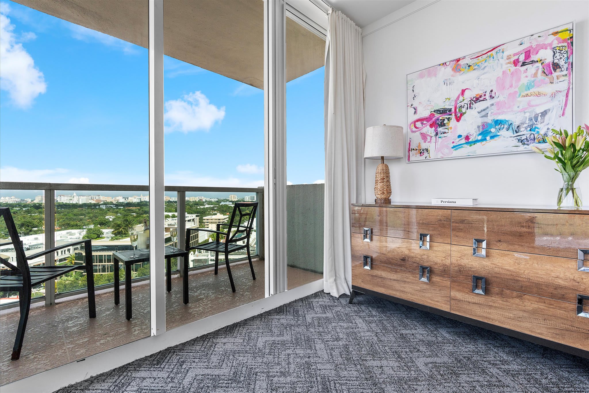 A corner of a room with a wooden dresser and abstract art, leading to a balcony through sliding glass doors. The balcony has outdoor seating and offers a panoramic city view.