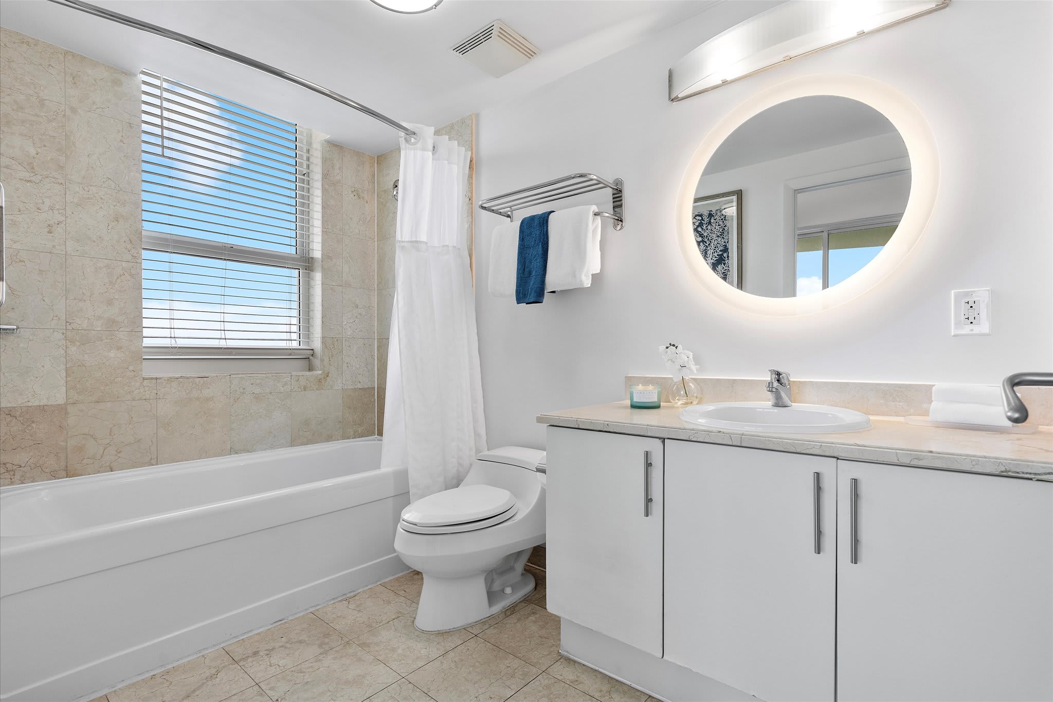 A clean bathroom with a white bathtub, toilet, and a vanity with a circular backlit mirror. The walls are tiled in a light beige stone, and a window with blinds offers natural light.