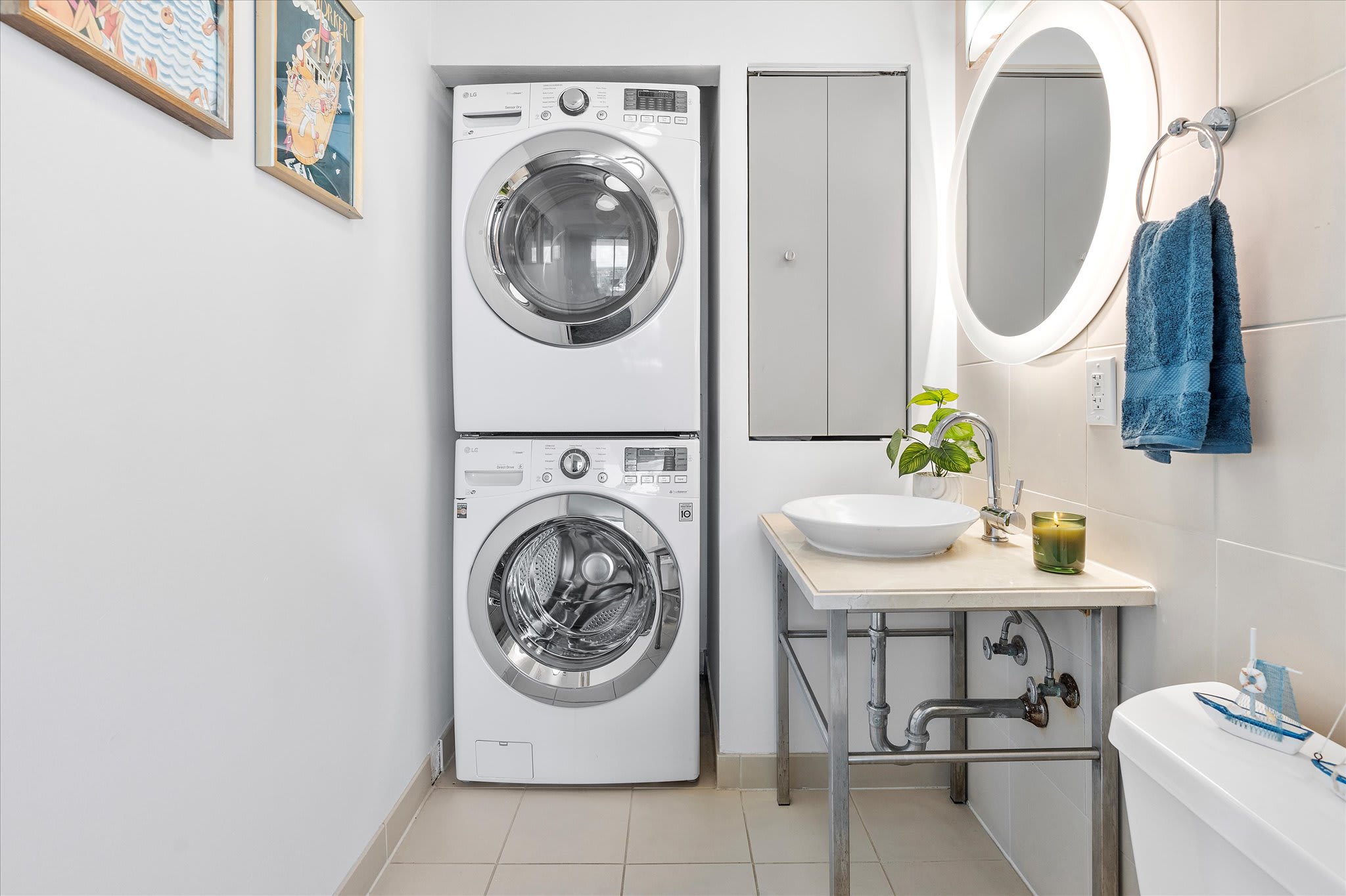 A compact utility area featuring a stacked washer and dryer unit, a small vanity with a vessel sink, and a toilet. A circular backlit mirror hangs above the sink.