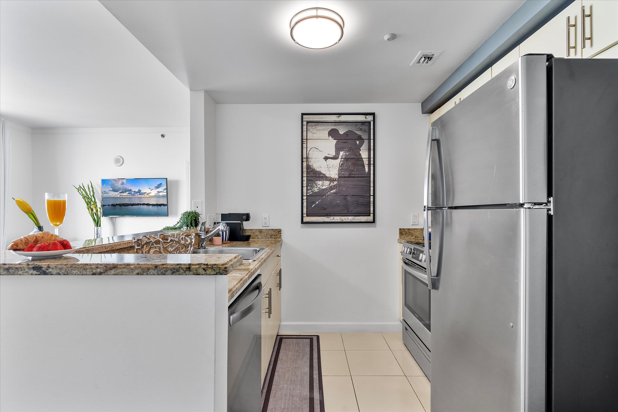 A modern kitchen features stainless steel appliances including a refrigerator and a range, light-colored cabinets, and granite countertops. A framed artwork hangs on the wall, and the floor is tiled.