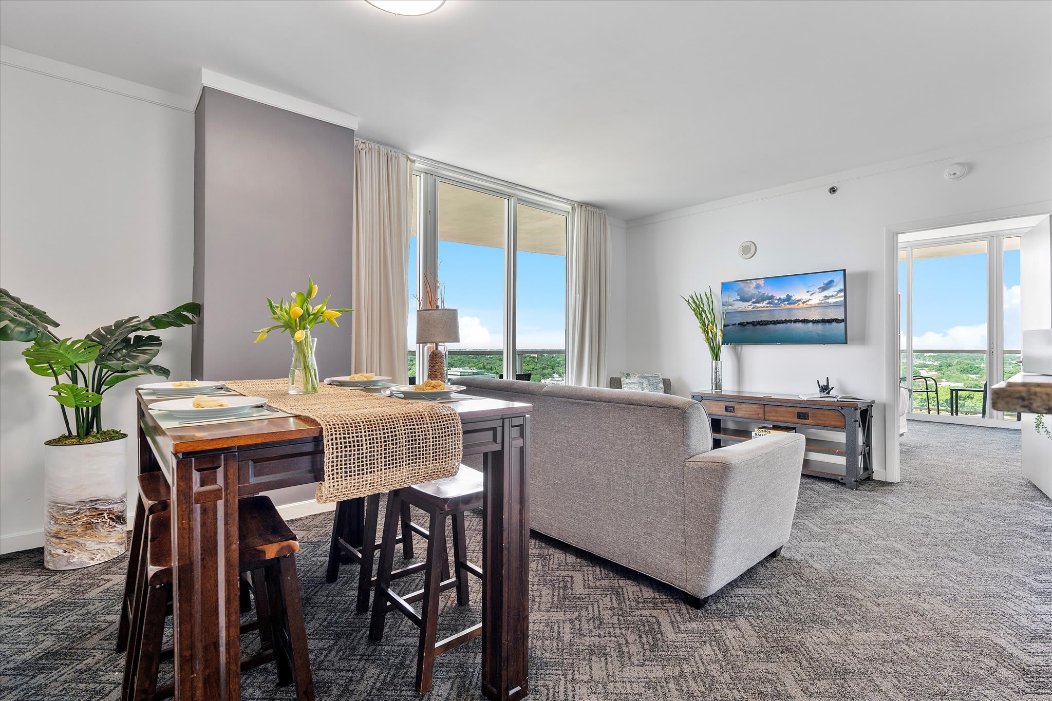 An open-concept living and dining area features a high-top wooden dining table with four stools, a light gray sofa, and floor-to-ceiling windows leading to a balcony with a view. The room is carpeted with white walls and a gray accent wall.