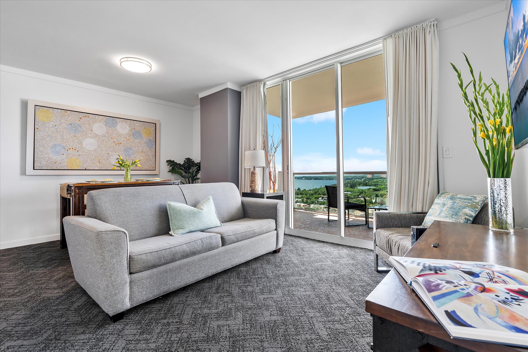 A cozy living room features a light gray sofa, a patterned armchair, and a wooden coffee table with magazines. Floor-to-ceiling windows offer a view of the bay and city, complemented by a large abstract painting above the sofa.
