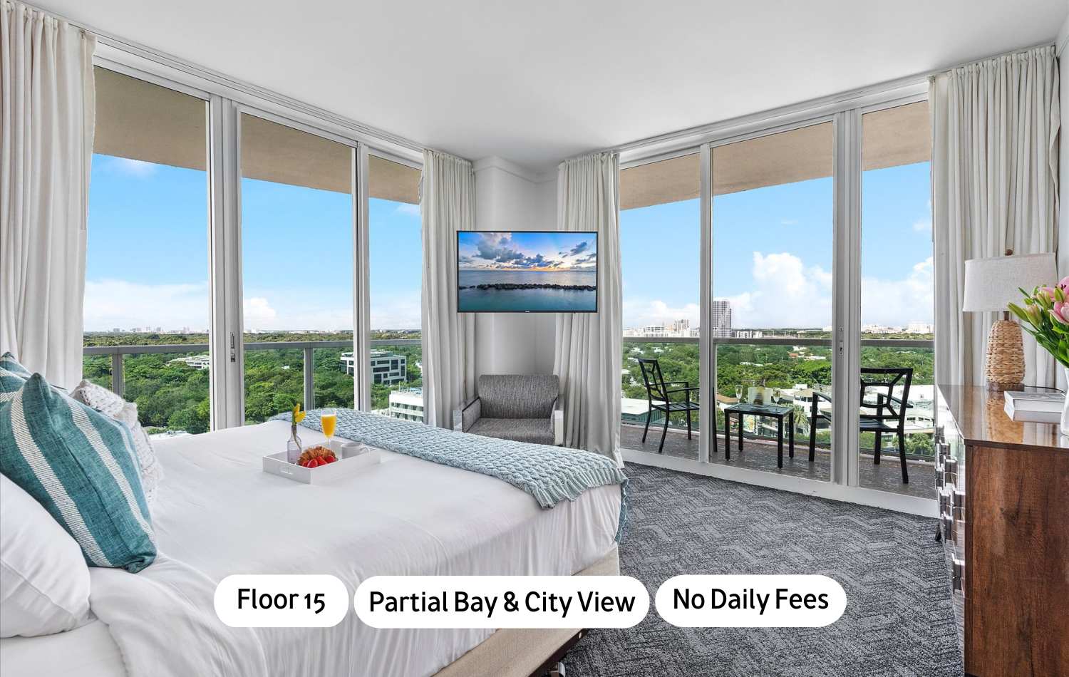 A bright bedroom features a white bed with blue and white bedding, a breakfast tray, and floor-to-ceiling glass doors leading to a balcony. A wall-mounted TV is visible, offering partial bay and city views.