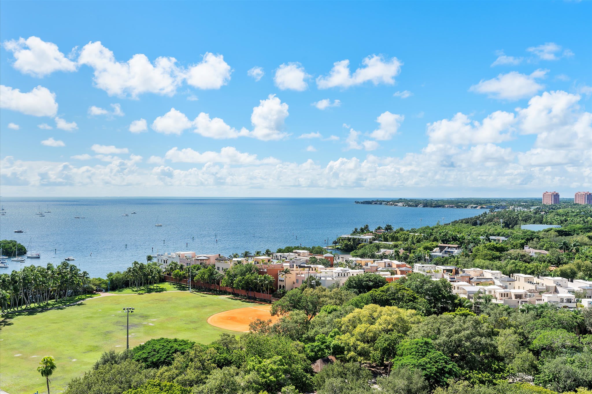 An expansive aerial view from a high floor showcases a wide bay dotted with boats, a vibrant green baseball field, and a residential area with various buildings under a bright blue sky with fluffy white clouds.