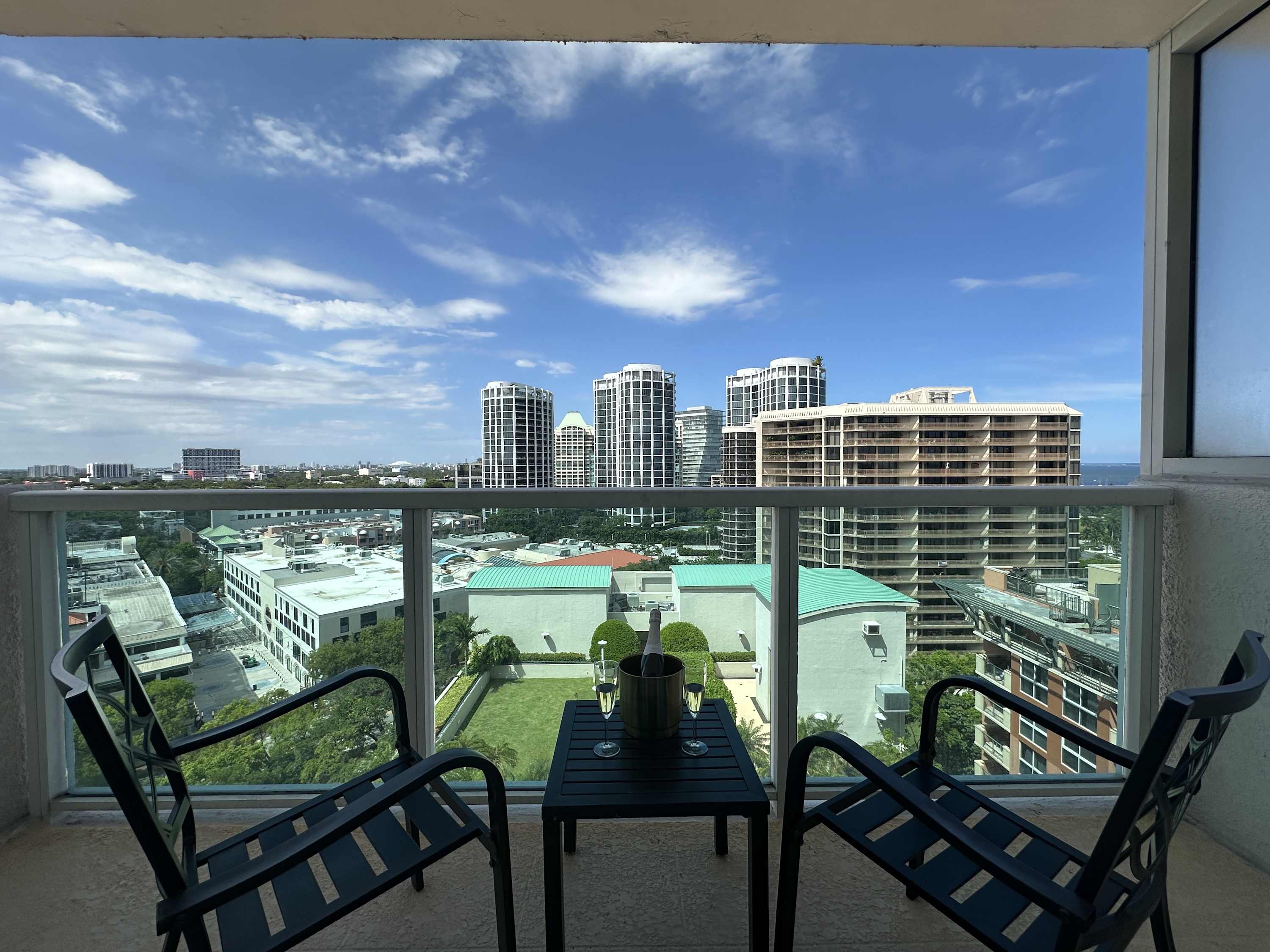 Private balcony with two black chairs and a small table holding an ice bucket with champagne, offering a clear view of multiple high-rise city buildings and green spaces below.