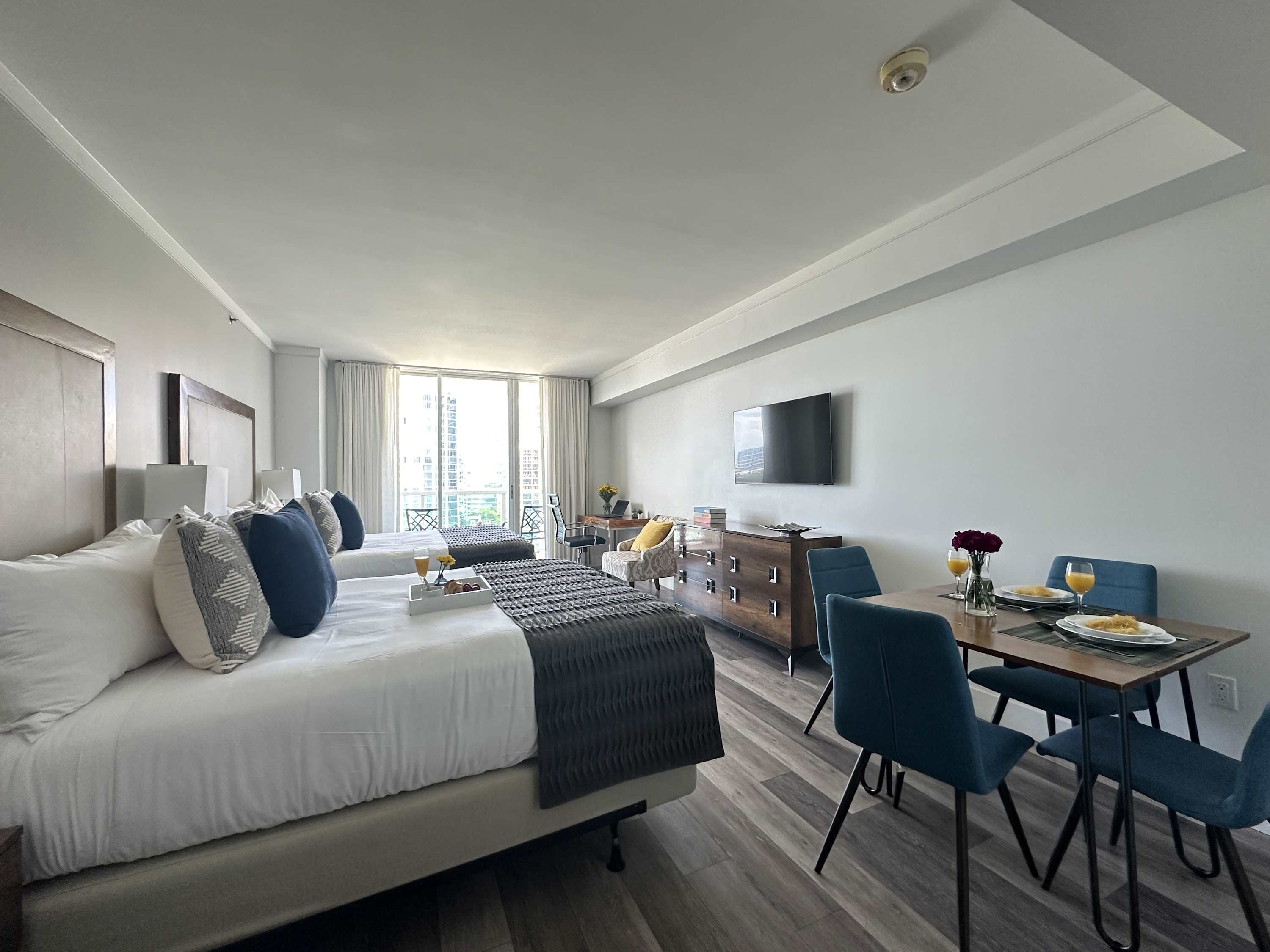 Expansive view of the studio apartment, showcasing a dining table with blue chairs, two queen beds, a dresser with a TV, and a bright balcony entrance, all on light wood-look flooring.