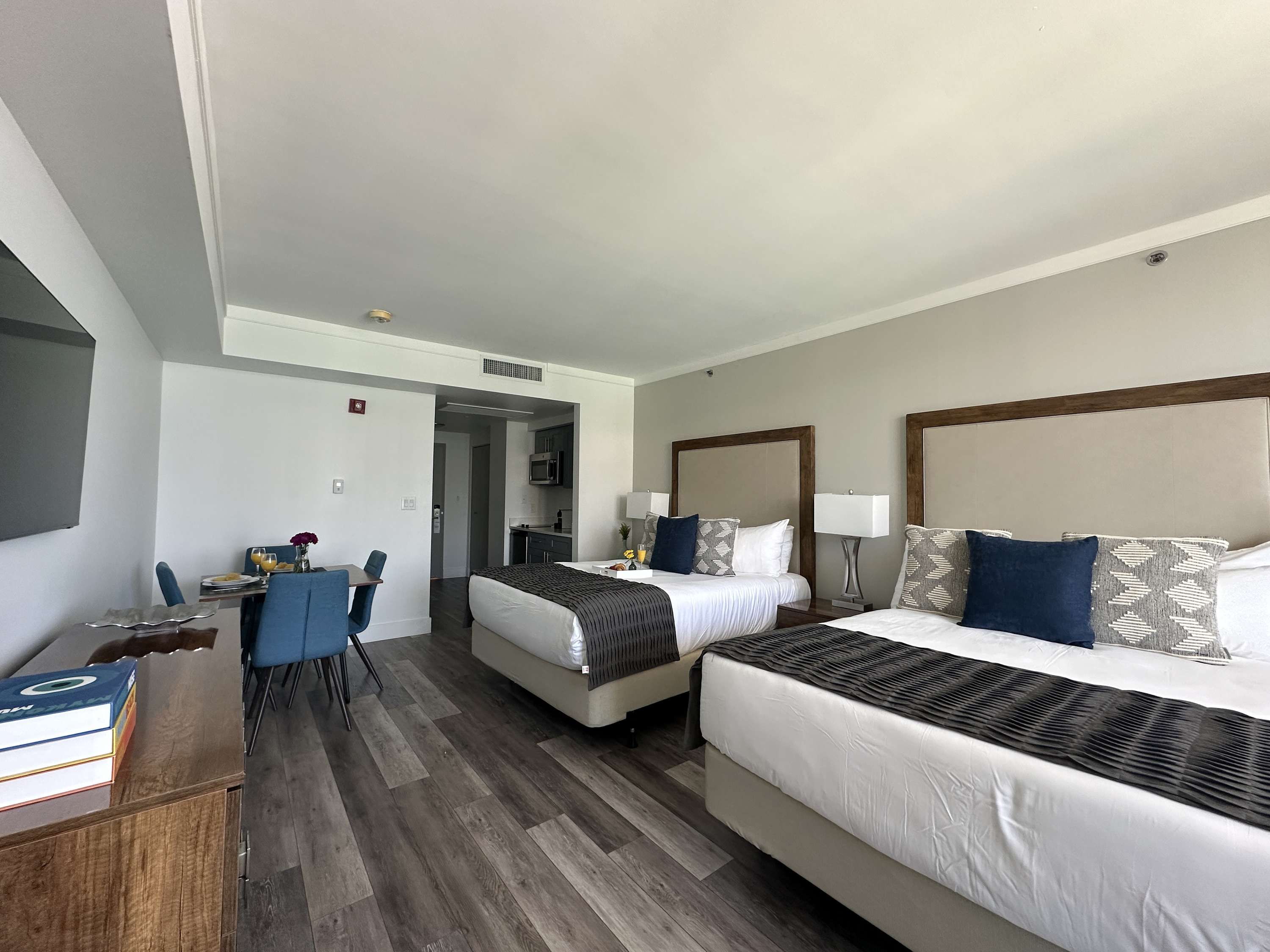 Bright studio apartment featuring two queen beds with grey throws, a small dining area with a wooden table and blue chairs, and a glimpse of a modern kitchen, all set on light wood-look flooring.