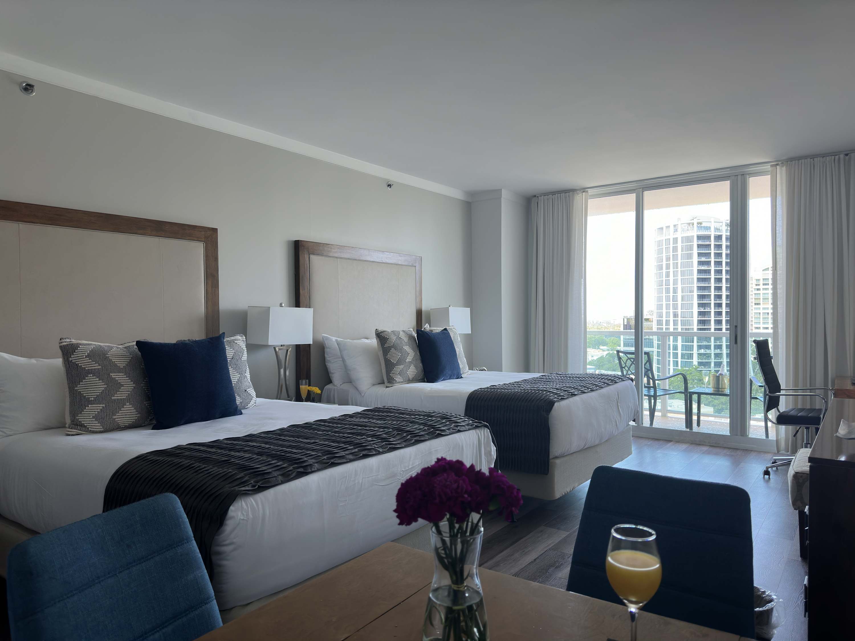 View from the dining area towards the sleeping space, showing two queen beds with grey throws and a large sliding glass door leading to a balcony with city views.