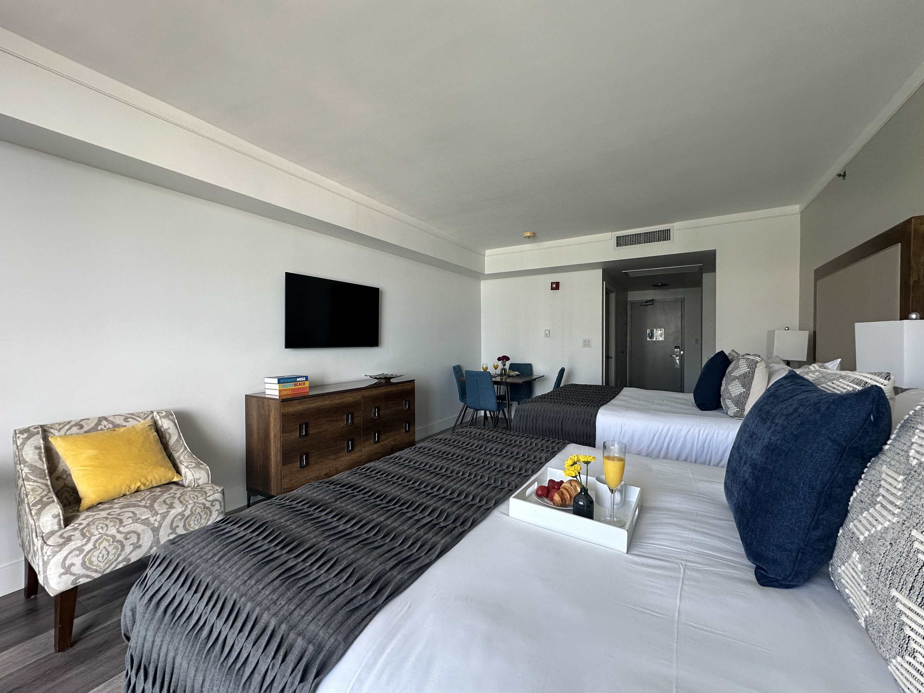 Spacious studio room with two queen beds, a long wooden dresser topped with a flat-screen TV, and a patterned armchair with a yellow pillow, all against light walls and wood-look flooring.