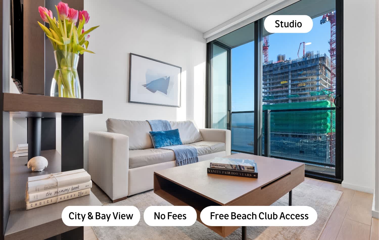 Modern living area with a light grey sofa, wooden coffee table, and a large window offering city and bay views, including a construction site. A dark wood shelving unit holds a vase of pink tulips.