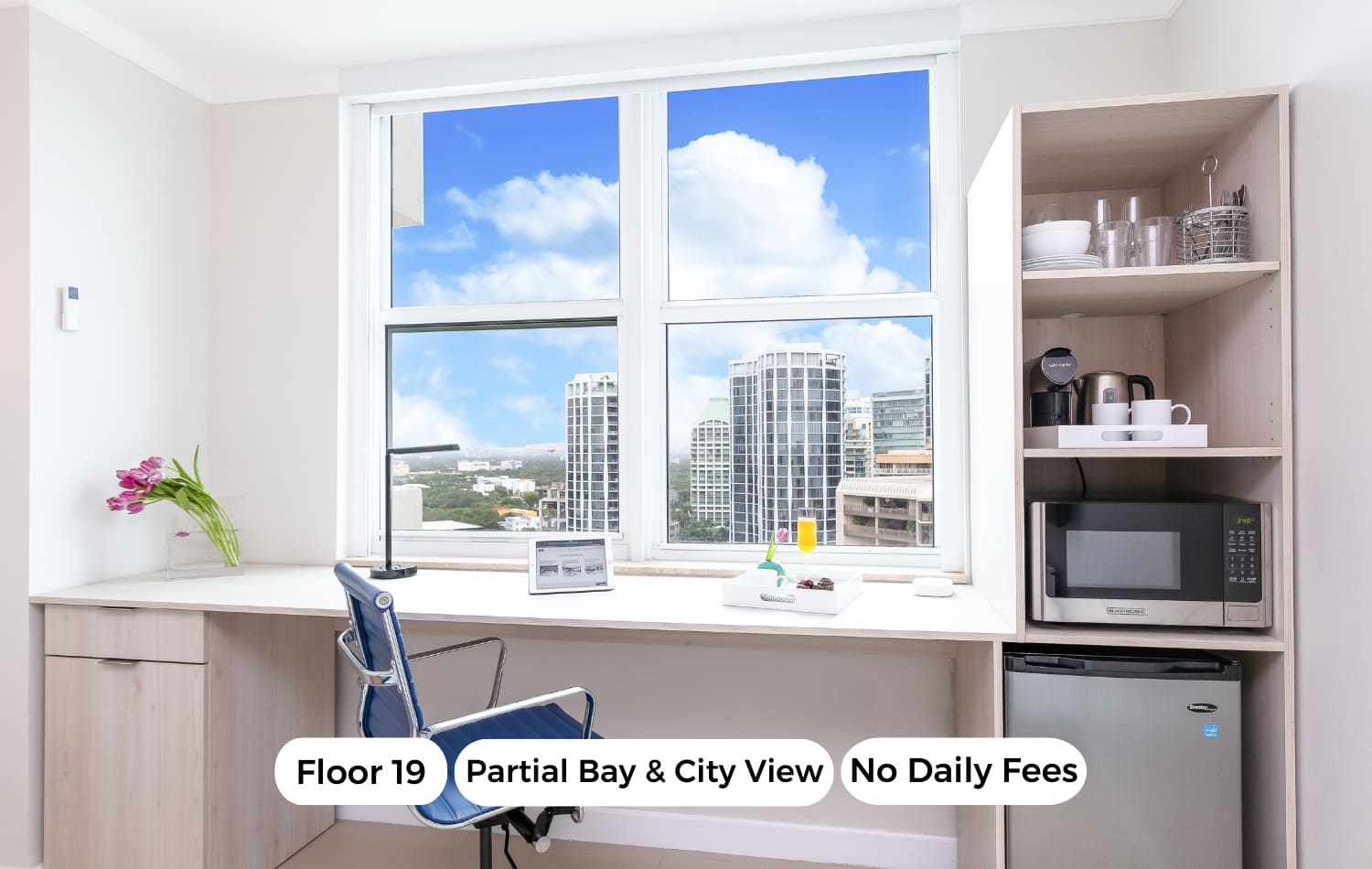 Bright workspace with a white desk, blue office chair, and large window offering a partial bay and city view, featuring a mini-fridge and microwave on the side.