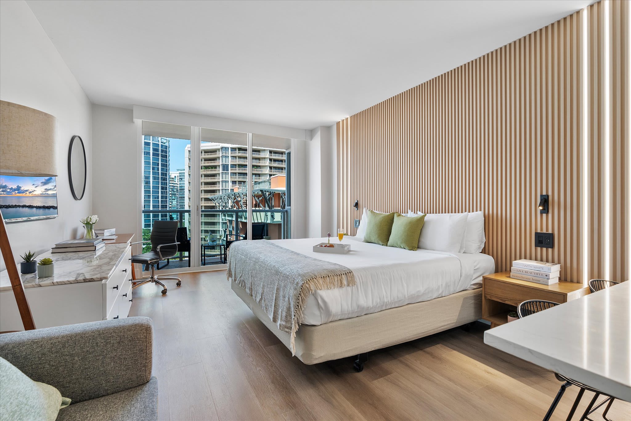 Bright studio bedroom with a queen-sized bed, a wooden slatted accent wall with integrated lighting, and floor-to-ceiling glass doors leading to a balcony. A desk with an office chair and a dresser with a TV are also visible.