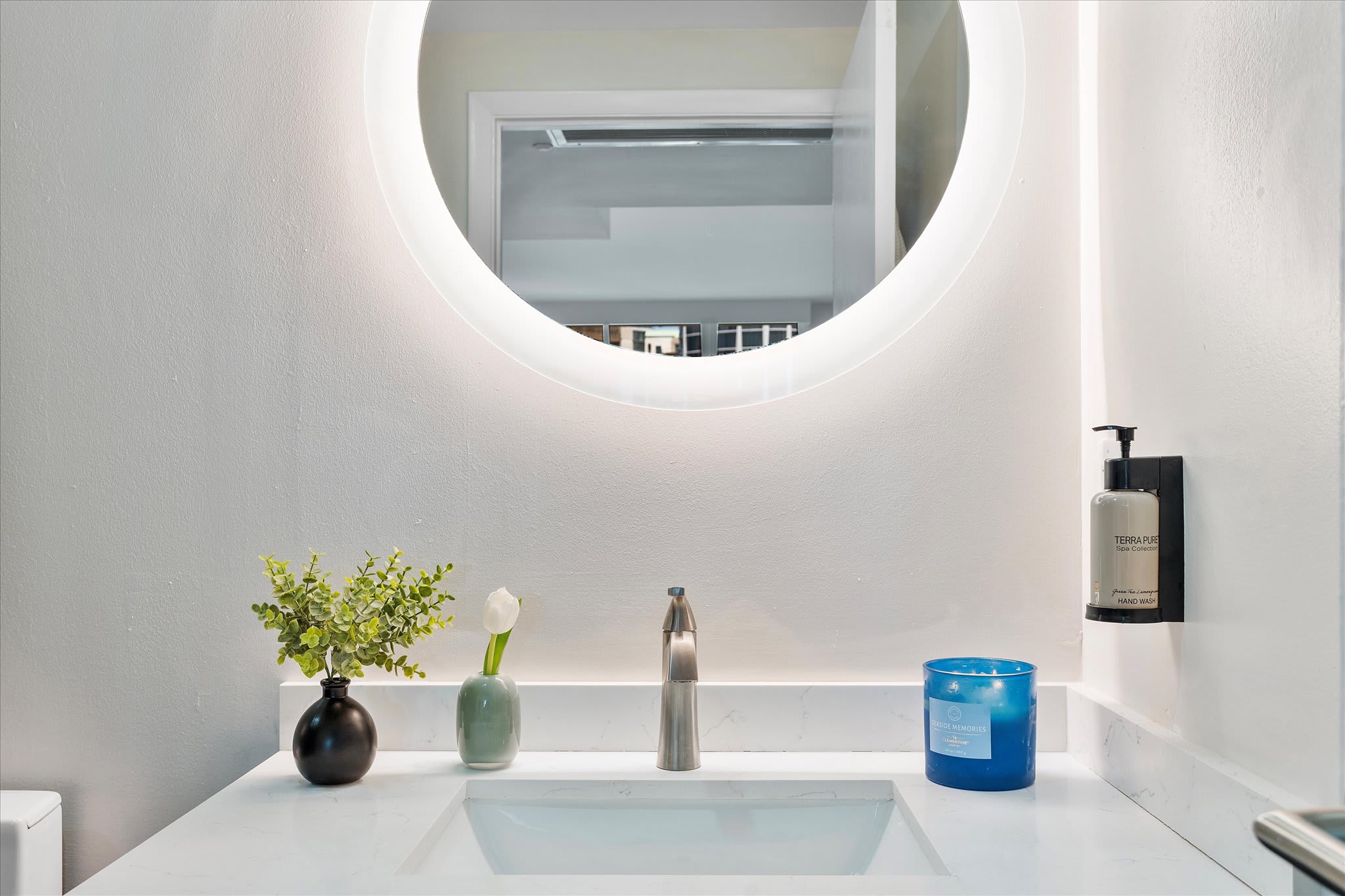 Close-up of a modern bathroom vanity featuring a white countertop, rectangular sink, and a sleek faucet. A large round backlit mirror illuminates the space, complemented by decorative accents and a wall-mounted soap dispenser.