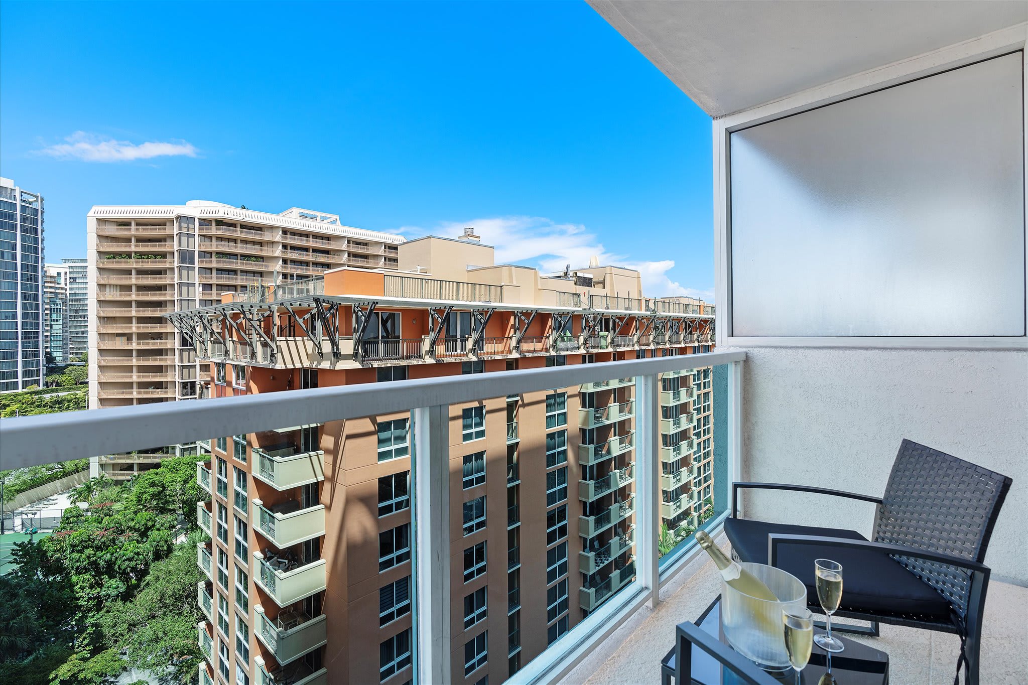 Balcony with glass railings, featuring two black chairs and a small table with a bottle of champagne and two glasses. The view shows surrounding high-rise buildings and a bright blue sky.