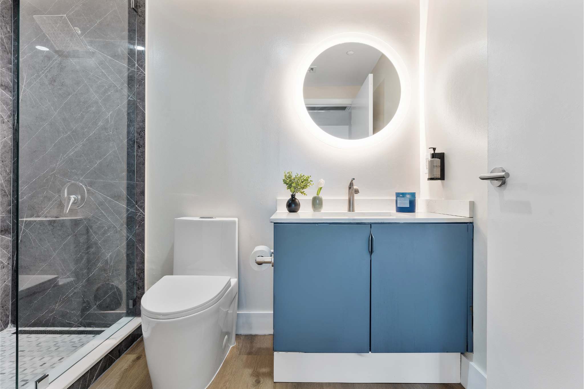 Contemporary bathroom with a white toilet, a blue vanity with a white countertop, and a striking round backlit mirror. A spacious glass-enclosed shower with dark gray marble-patterned walls is visible to the left.