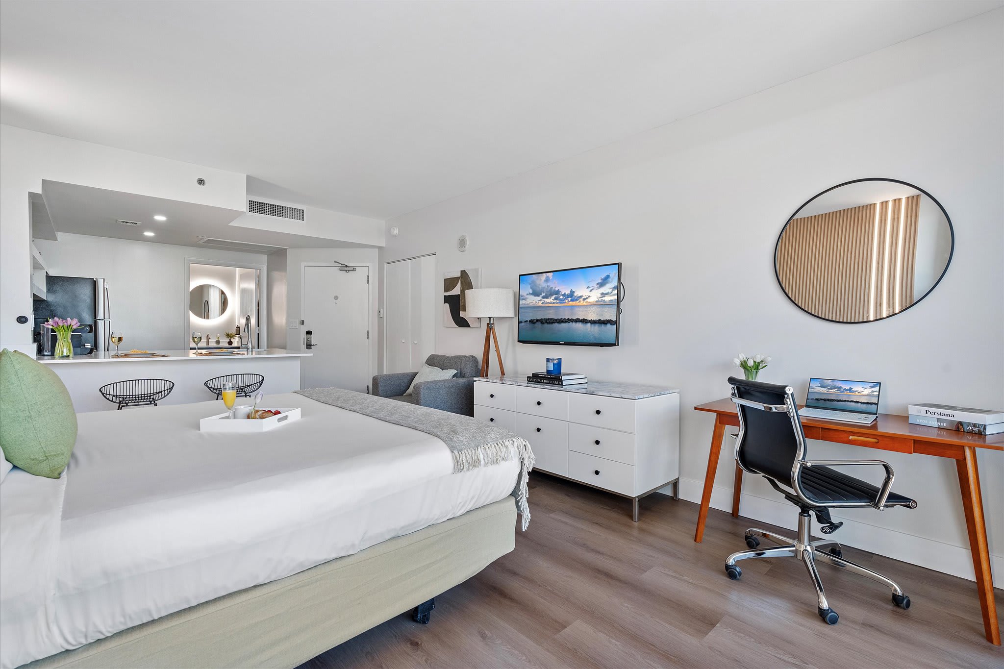 Full view of the open-concept studio, showing the queen bed, a small gray armchair, a white dresser with a TV, and a wooden desk. The kitchen area with a breakfast bar and a glimpse of the bathroom are visible in the background.