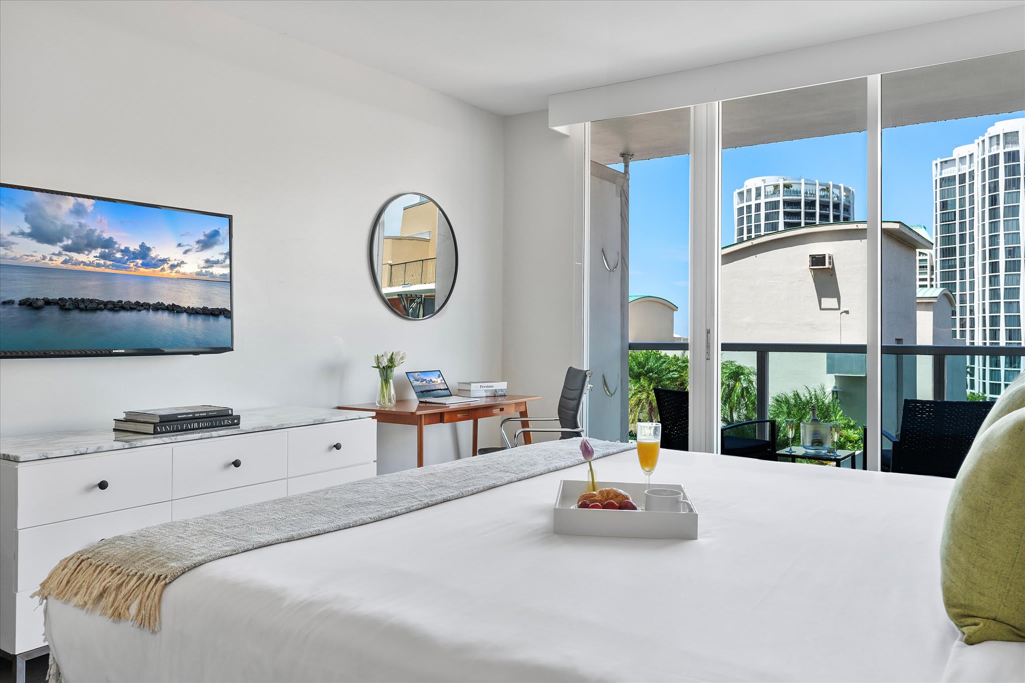 View of the studio bedroom from the bed, showing a white dresser with a flat-screen TV, a wooden desk with a laptop, and a round mirror. Floor-to-ceiling glass doors open to a balcony with city views.