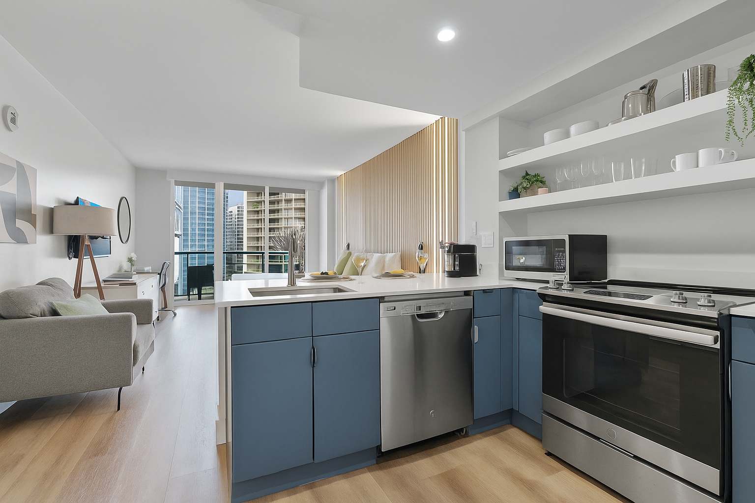 Modern open-concept kitchen featuring blue flat-panel cabinetry, white quartz countertops, stainless steel appliances, and open shelving. It seamlessly connects to a bright living area with a sofa and city views through large windows.