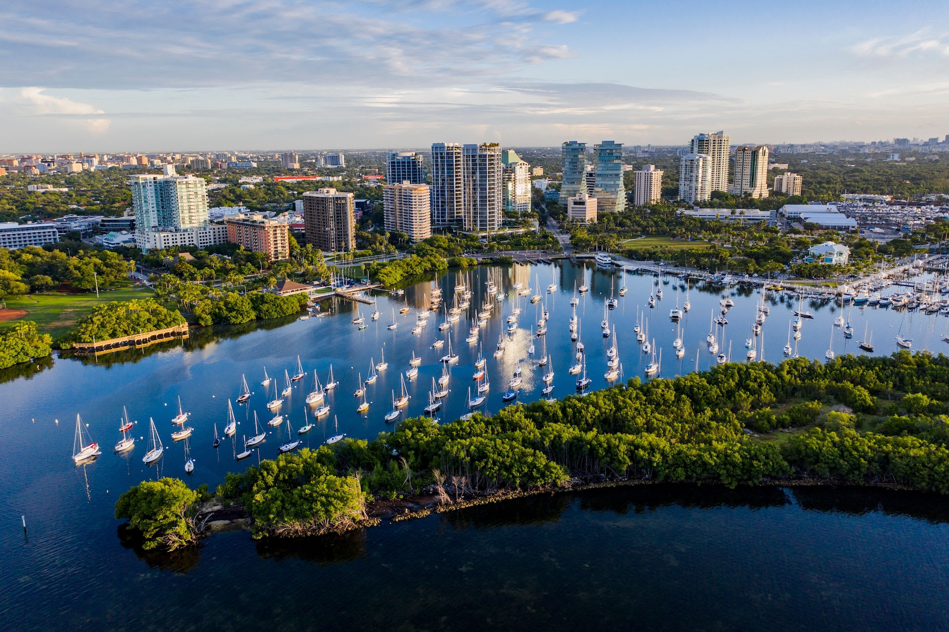 Stunning aerial view of a vibrant city skyline featuring numerous high-rise buildings, a serene bay dotted with sailboats, and lush green waterfront areas under a partly cloudy sky.