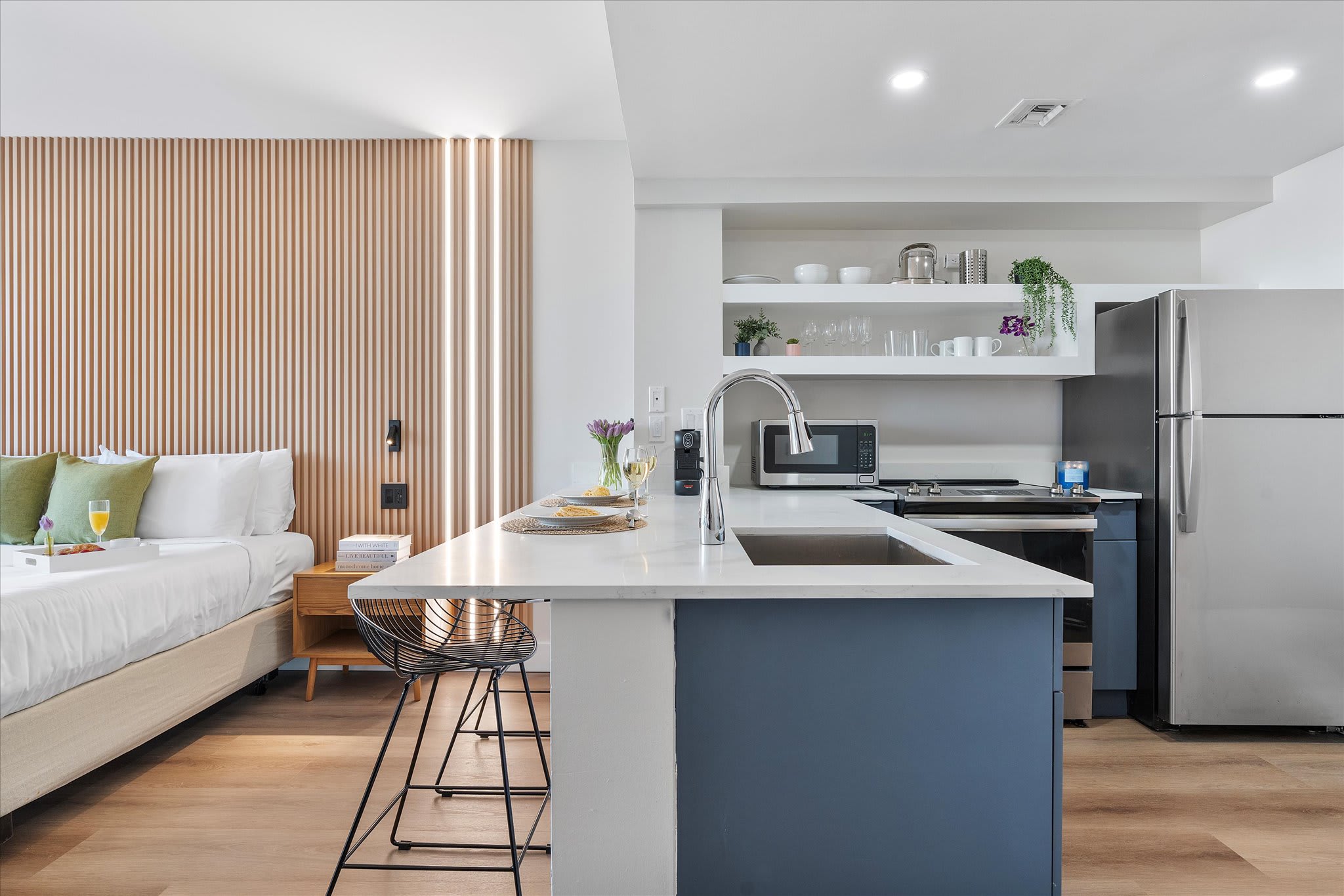 Modern kitchen area with a white quartz island featuring a sink and two bar stools, integrated into an open-concept studio space. Stainless steel appliances, white floating shelves, and a wooden slatted accent wall are visible.