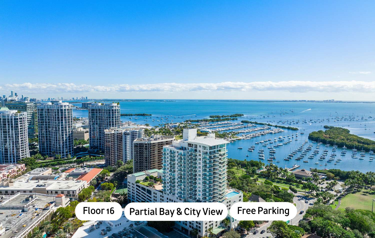 High-angle aerial view of a coastal city with numerous high-rise buildings, a large bay filled with boats and marinas, and a clear blue sky. Text overlays indicate "Floor 16", "Partial Bay & City View", and "Free Parking".