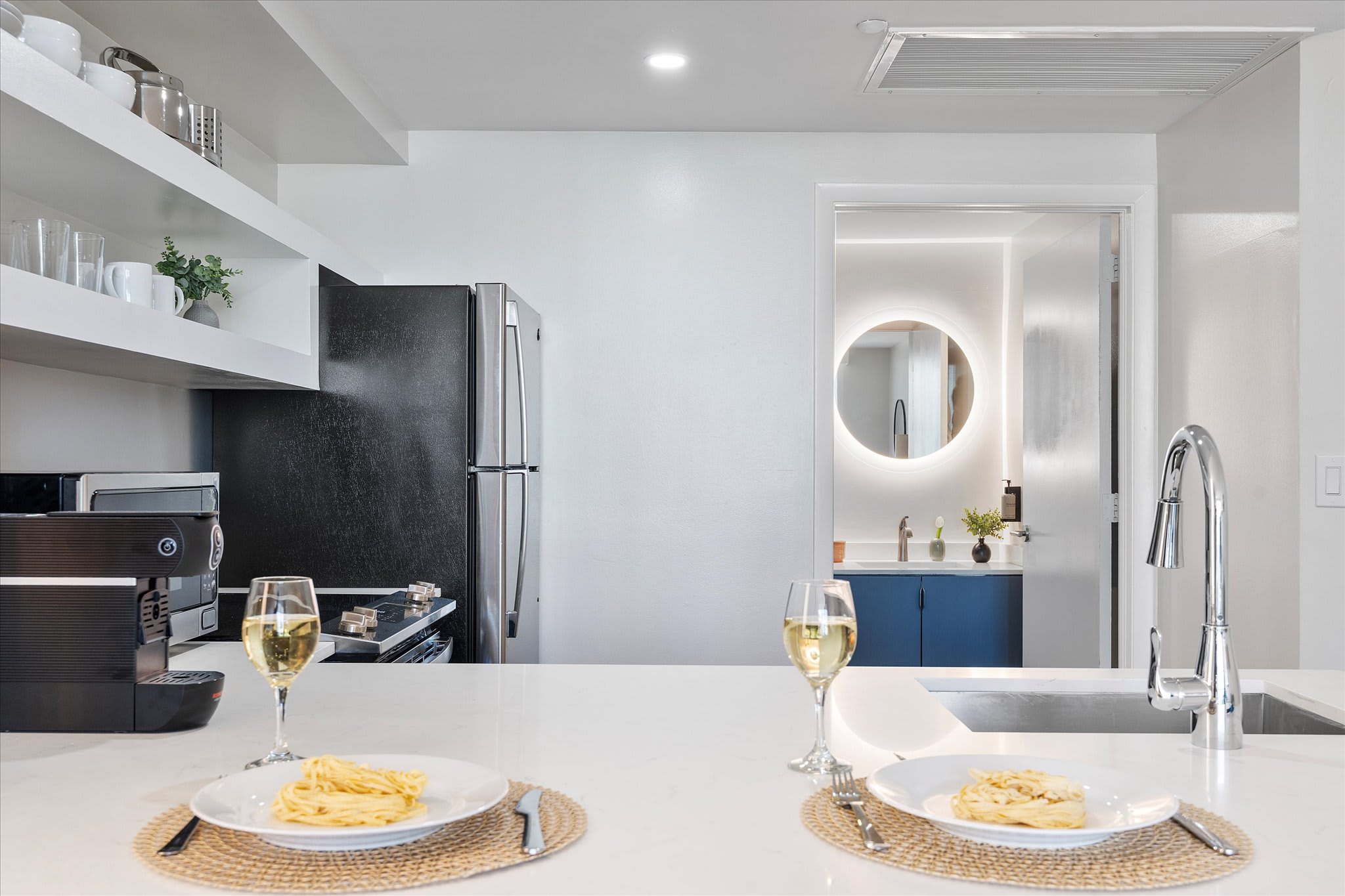 View from the kitchen island showing two plates of pasta and wine glasses, with a modern kitchen featuring a black refrigerator and a glimpse of a bathroom with a circular backlit mirror in the background.