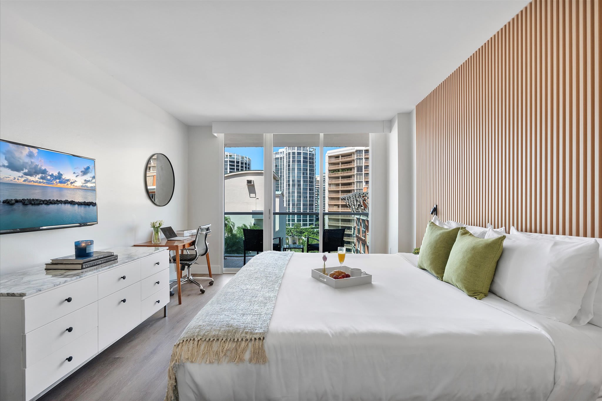 Wide shot of the studio bedroom featuring a queen bed with green pillows, a wooden slatted accent wall, and floor-to-ceiling glass doors leading to a balcony with city views. A white dresser with a TV and a desk are on the left.