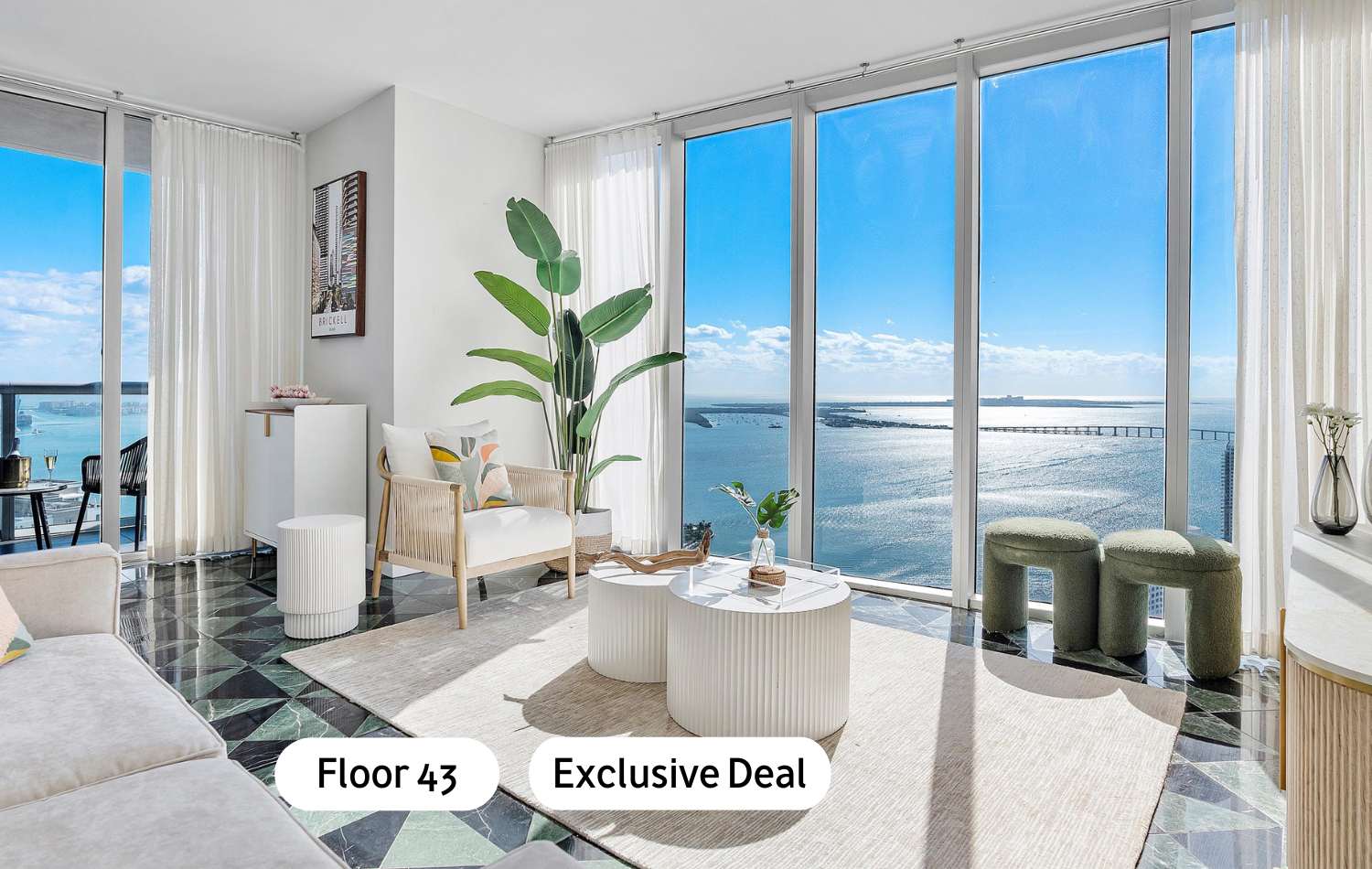 Bright living room with floor-to-ceiling windows offering expansive ocean views, featuring a light-colored sofa, two round white coffee tables, and a large green plant, all set on a patterned dark green and black tile floor.