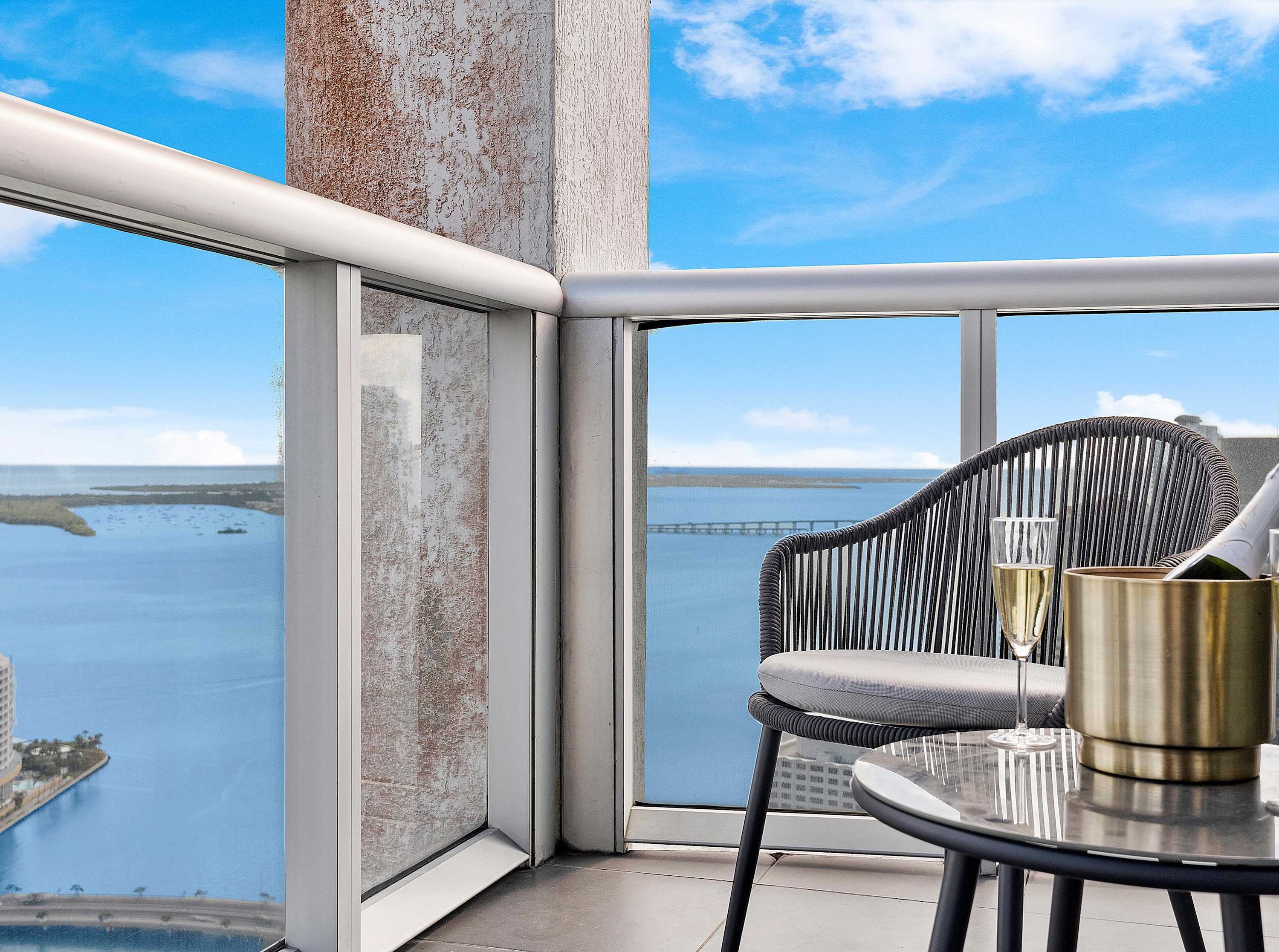 Close-up of a balcony corner with a stylish grey woven chair, a small round table, and a glass of champagne, overlooking a vast expanse of blue water and distant bridges.