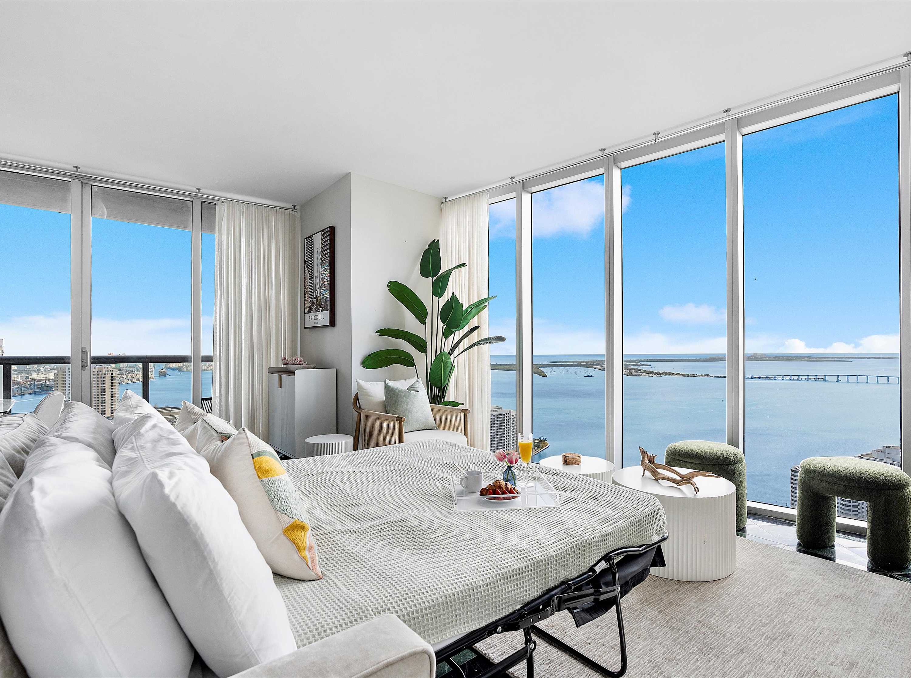 Spacious living room with a pull-out sofa bed, floor-to-ceiling windows offering panoramic ocean and city views, light-colored rug, and minimalist white side tables.
