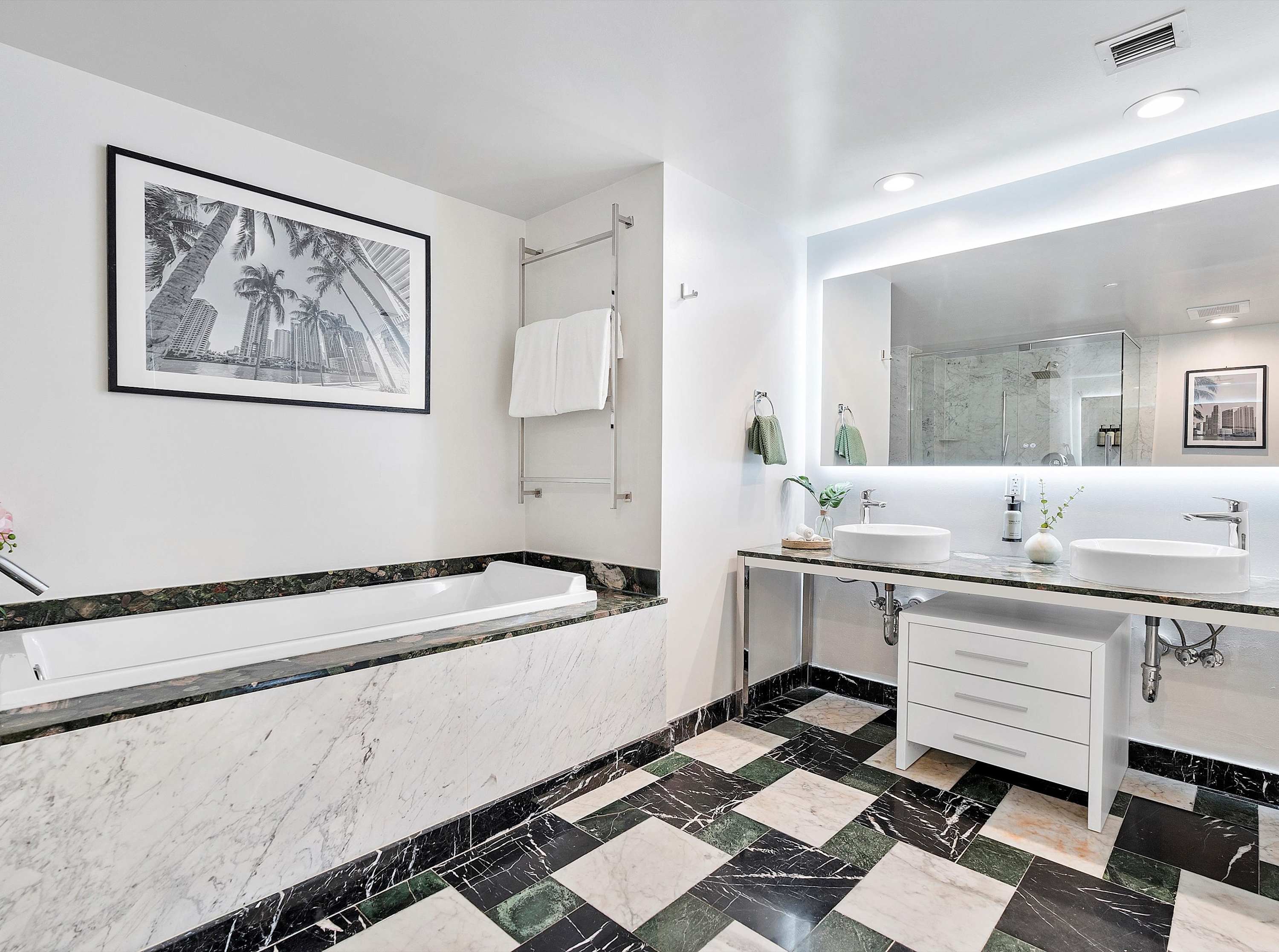 Modern bathroom with a large built-in bathtub, a double vanity with white vessel sinks, and a large illuminated mirror, set against a striking black and green geometric tile floor and white marble accents.