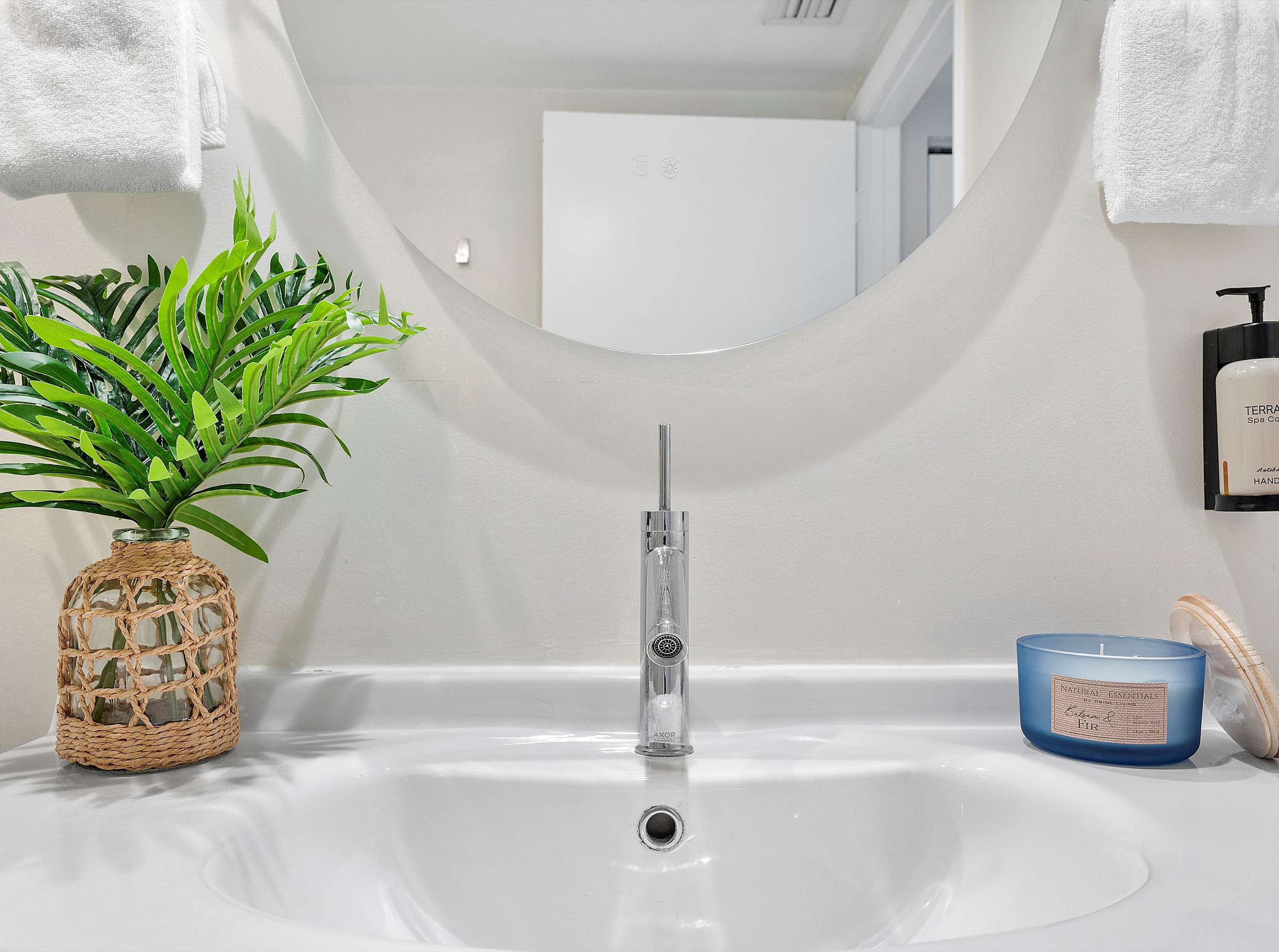 Close-up of a sleek white bathroom sink with a modern chrome faucet, a round mirror reflecting the room, and a small plant in a woven vase, alongside a blue candle and toiletries.