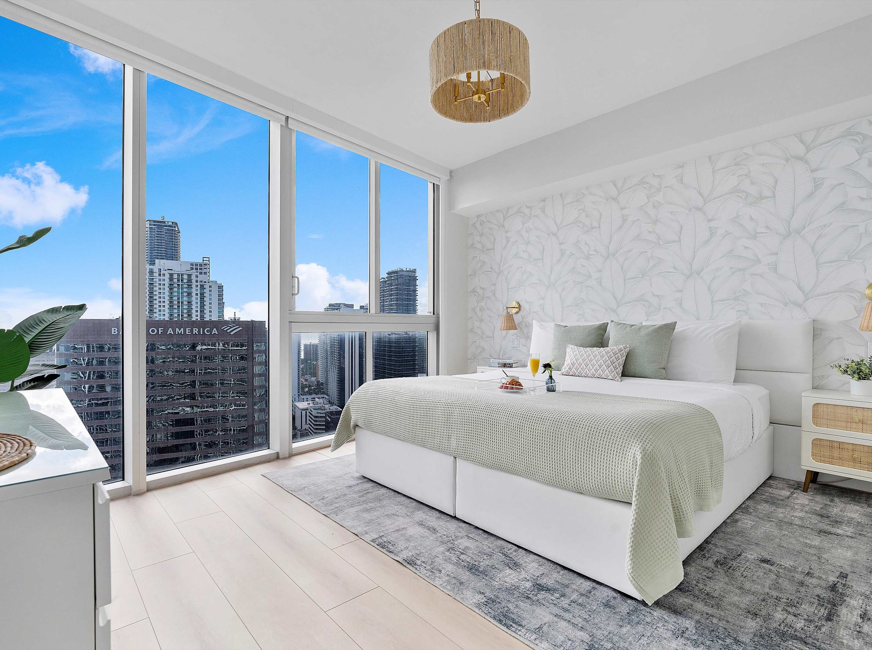 Bright bedroom with a large white bed, light wood flooring, and floor-to-ceiling windows offering a city view, featuring a decorative wallpaper accent wall behind the bed and a woven pendant light fixture.