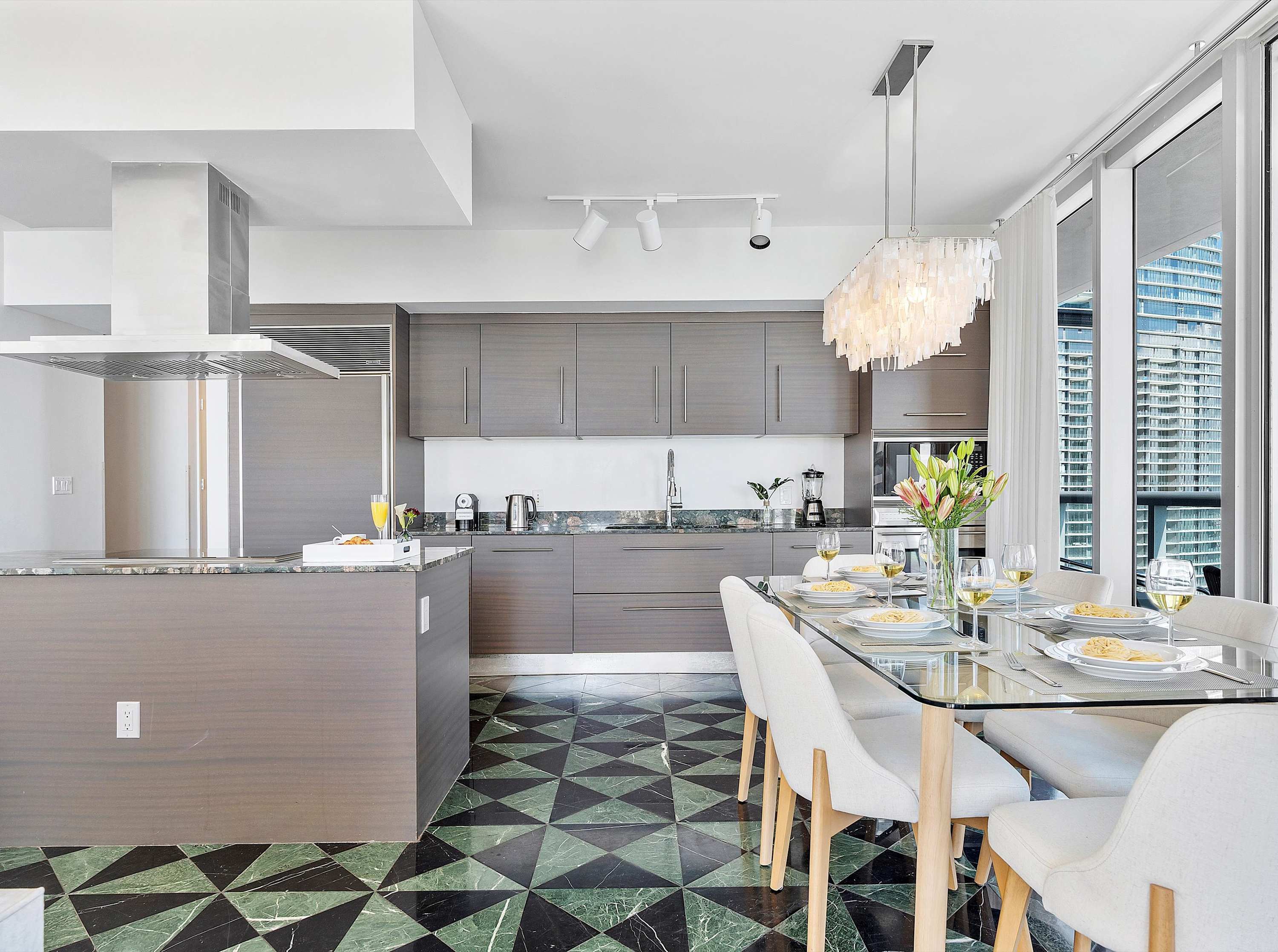 Modern kitchen with sleek gray cabinetry, stainless steel appliances including a built-in refrigerator and oven, and a glass dining table set for a meal, all illuminated by track lighting and a decorative chandelier, on a green and black geometric tile floor.