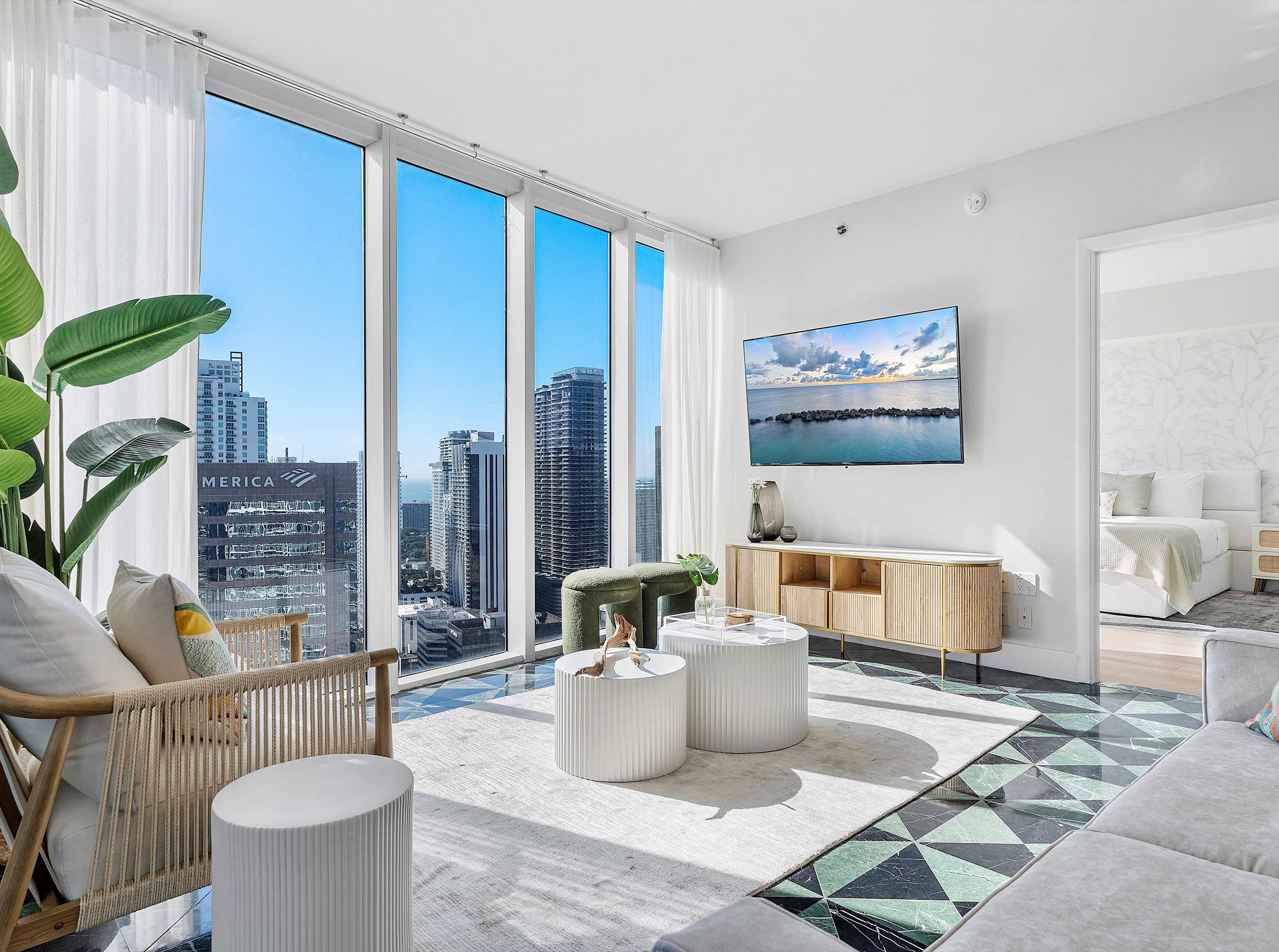 Bright living room with floor-to-ceiling windows showcasing a city skyline view, featuring a light-colored sofa, a woven armchair, two round white coffee tables, and a wooden media console with a mounted TV, all on a patterned dark green and black tile floor.
