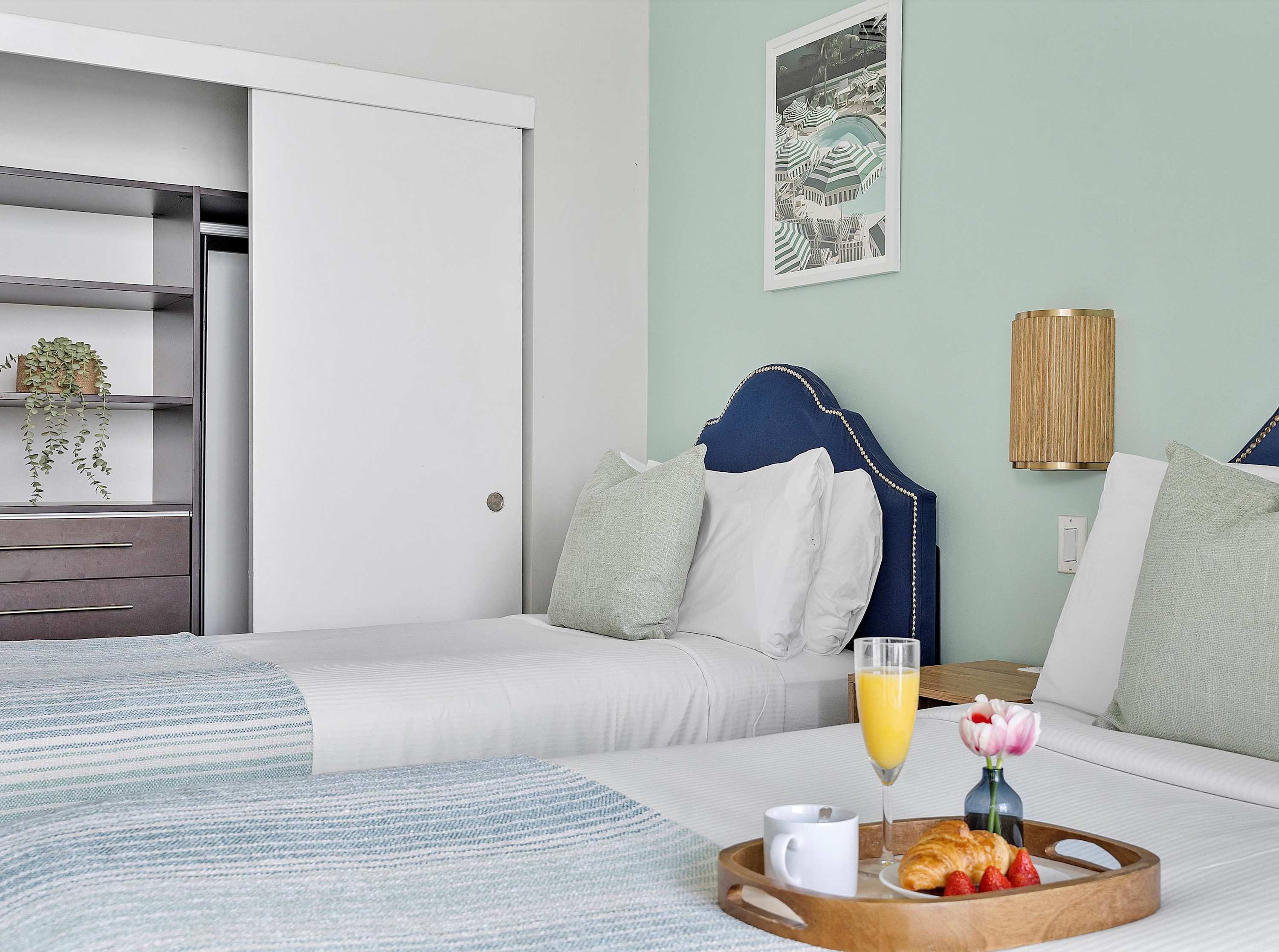 Cozy bedroom featuring two twin beds with blue upholstered headboards, light green pillows, a light blue and white striped throw, and a light mint green accent wall with a framed print.