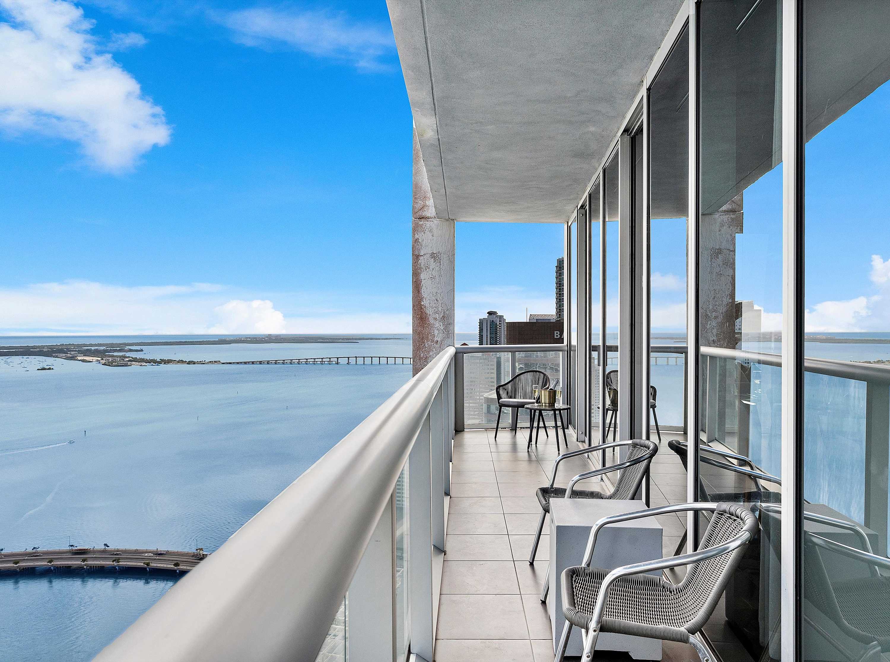 Spacious balcony with outdoor seating, offering panoramic views of the ocean and distant bridges under a clear blue sky, enclosed by a glass and metal railing.
