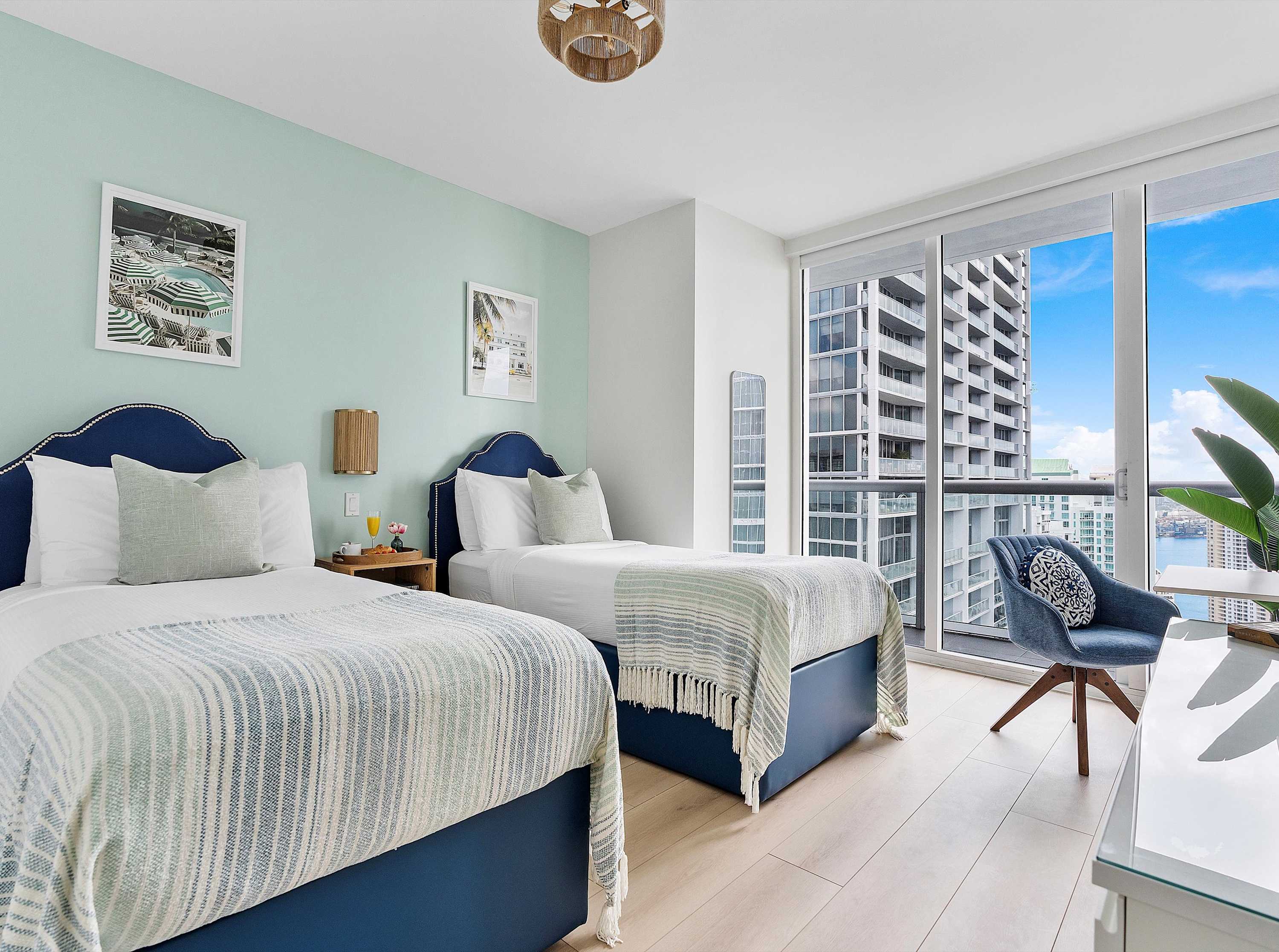 Serene second bedroom with two twin beds featuring blue upholstered headboards, a light blue accent wall, and floor-to-ceiling windows offering city and water views.