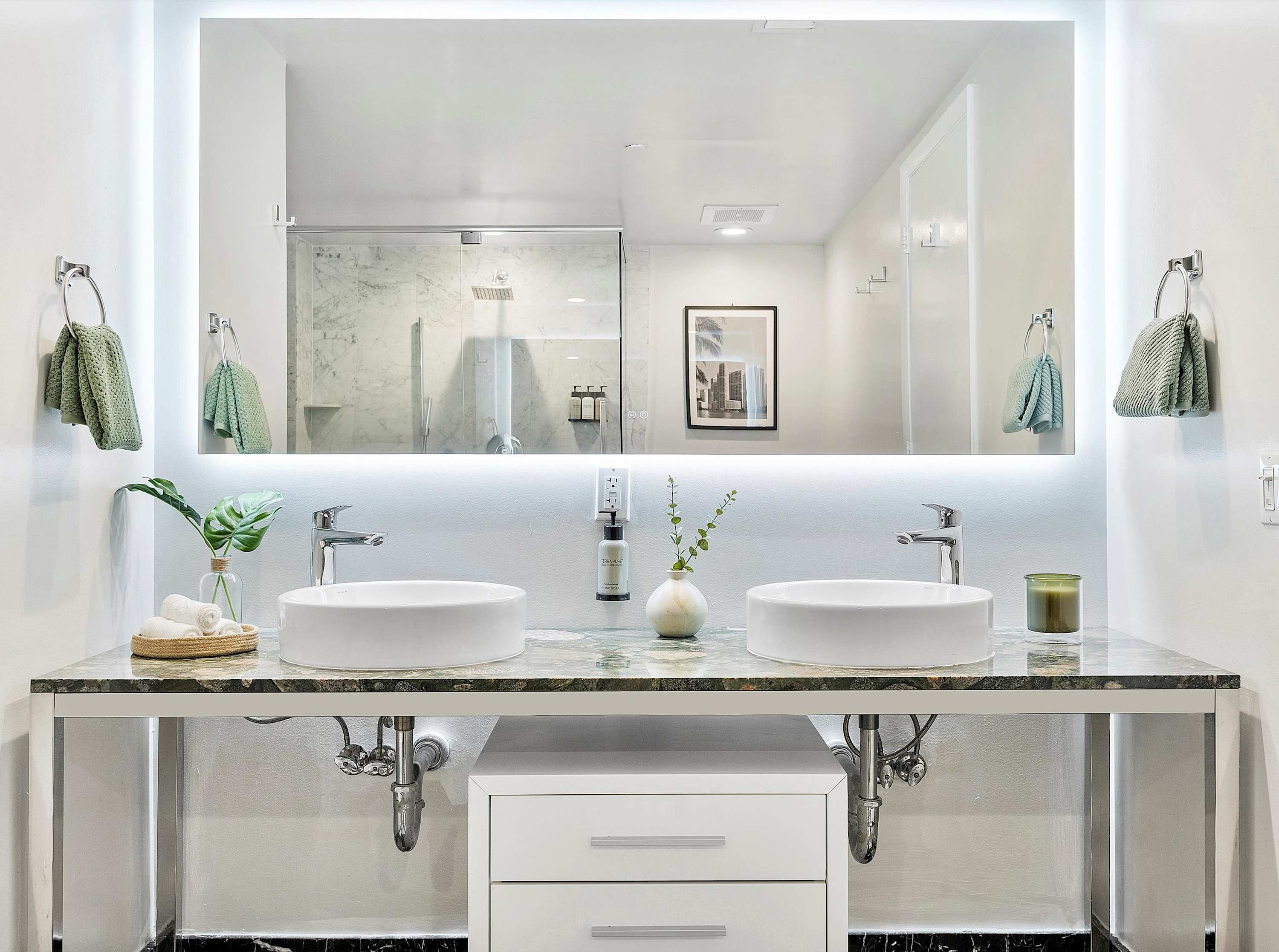 Modern bathroom with a double vanity featuring two vessel sinks, a green marble countertop, and a large illuminated mirror, reflecting a glass-enclosed shower.